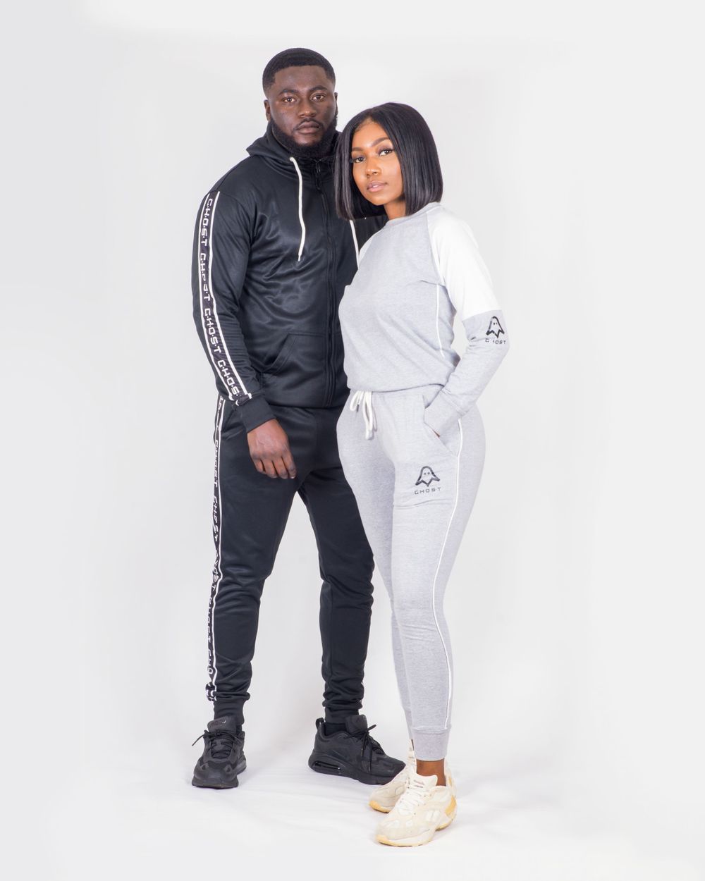 A Black man and woman posing together wearing matching Ghost brand tracksuits in black and grey colorways.