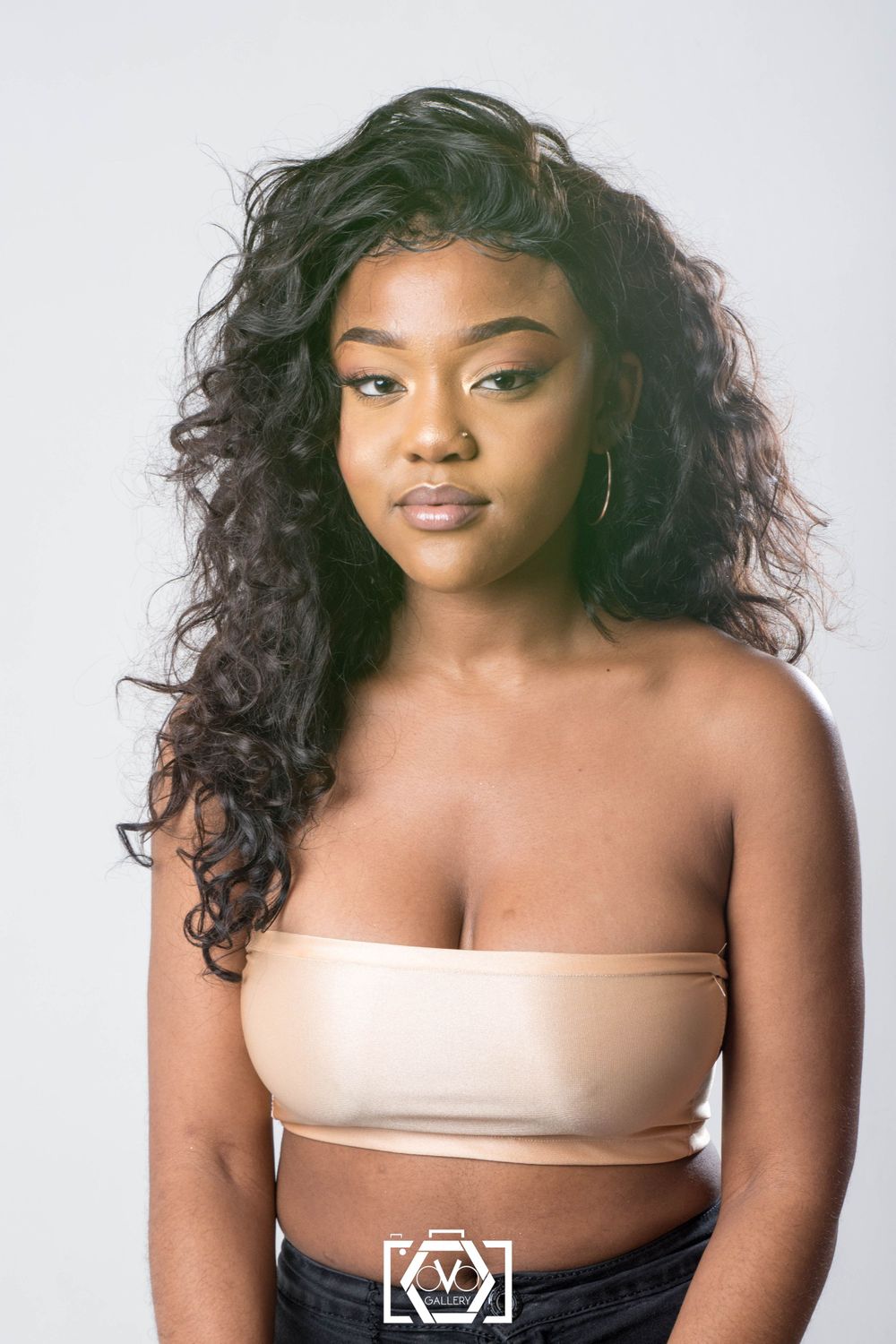Young Black woman with long curly hair wearing a nude bandeau top posing against a white background.