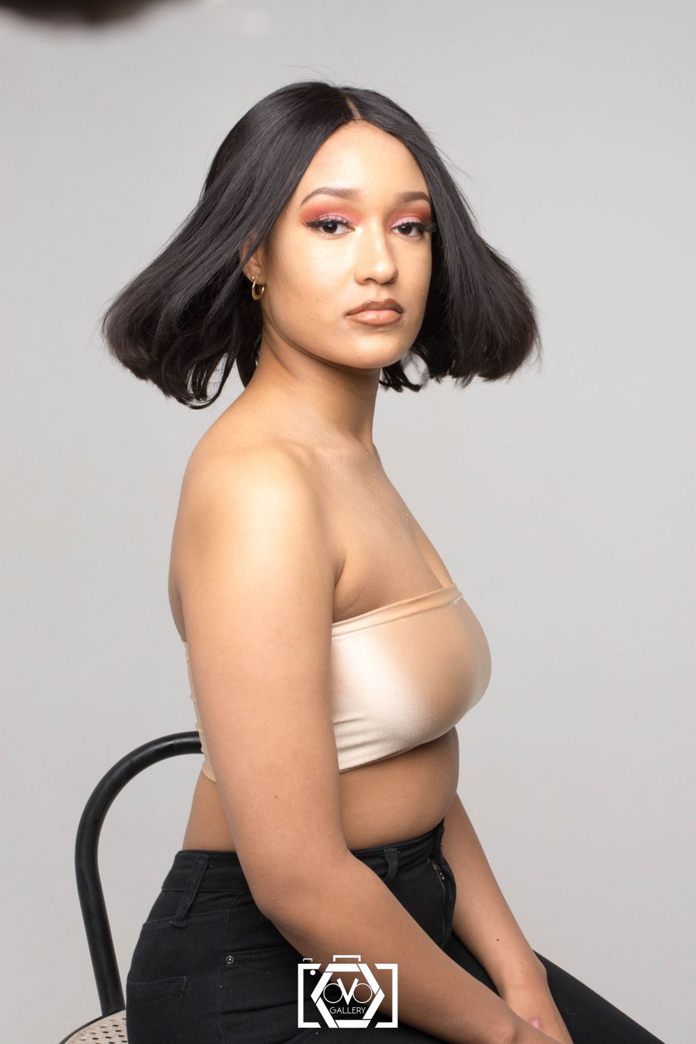 Young Black woman with a short bob hairstyle and pink eye makeup sitting on a chair in a studio.
