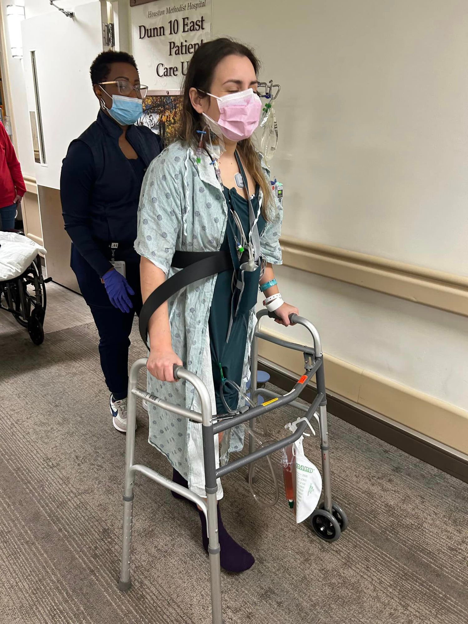 a brunette woman wearing a hospital gown and mask uses a walker in the hospital hallway