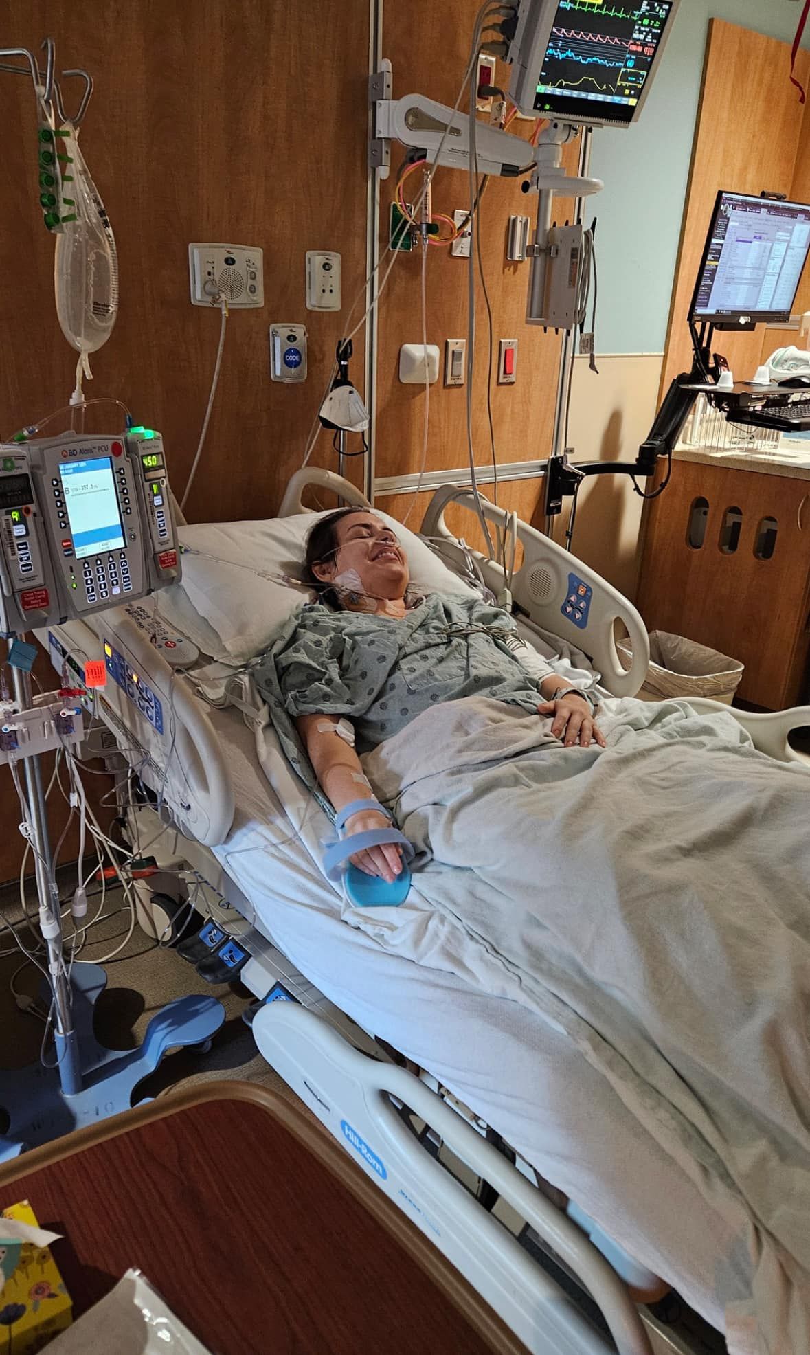 A brunette woman lies in a hospital bed smiling while covered in monitors and wires with blanket over her