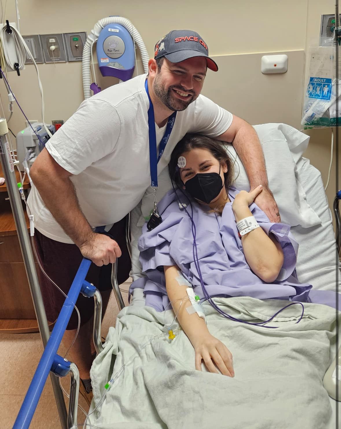 a patient sits up in her hospital bed with her husband standing nearby. His arm is around her and she holds his hand