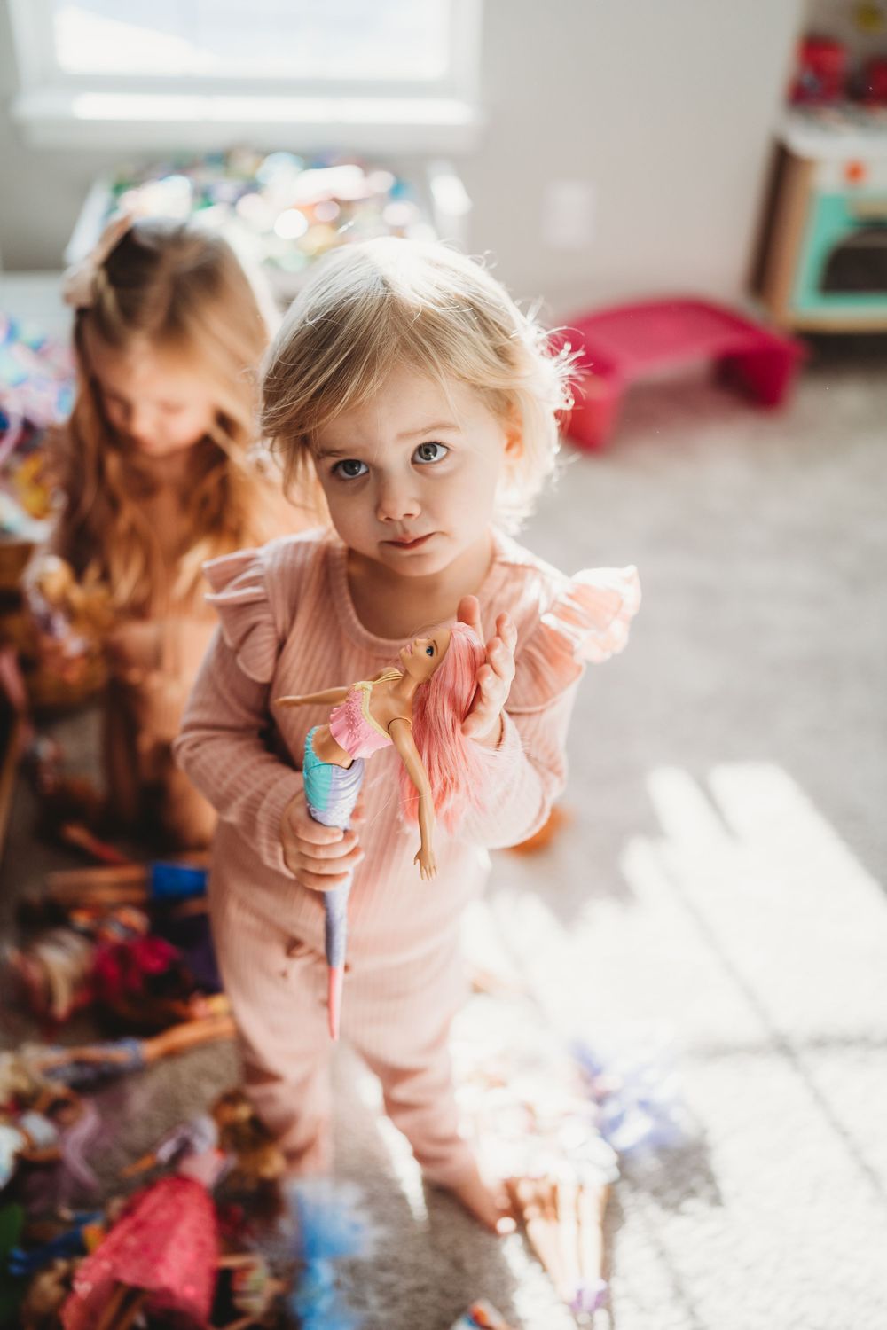 Child in pink dress playing in a sunlit room with toys scattered around.