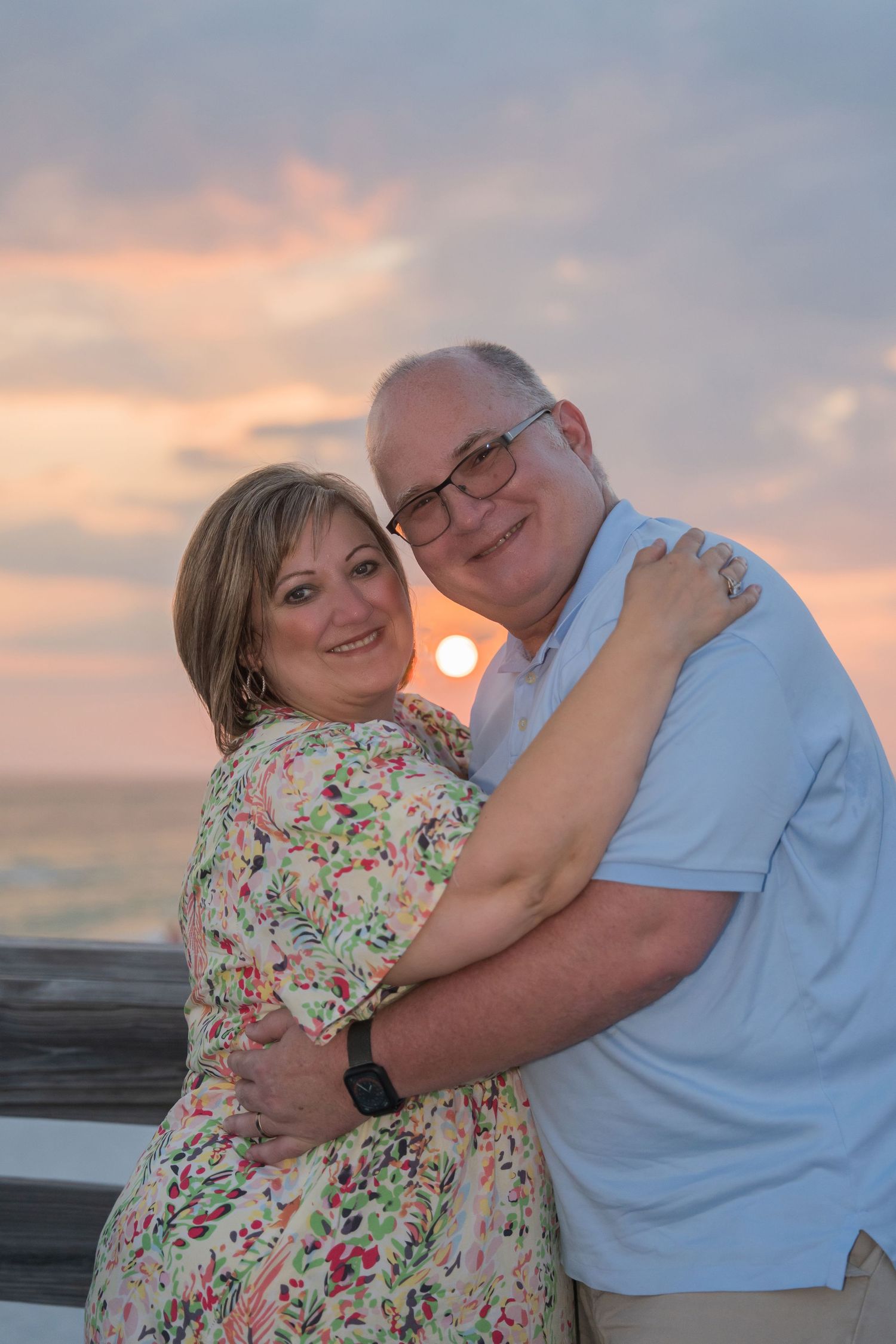 Couple embracing and smiling at sunset on a beach pier with colorful pink and orange sky.