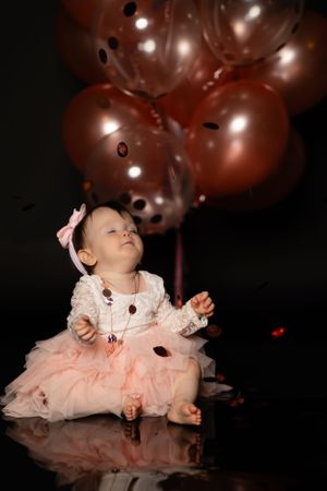 Baby in pink dress with bow looking up at floating balloons in dark studio setting.