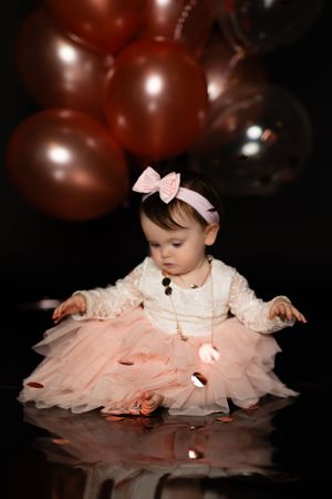 Baby in pink tutu dress with bow sitting on reflective floor with balloons in background.