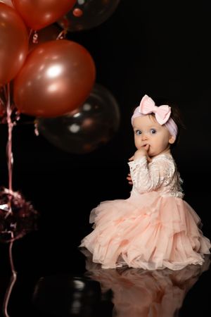 Baby in lace dress and pink bow posing with red balloons in dark studio setting.