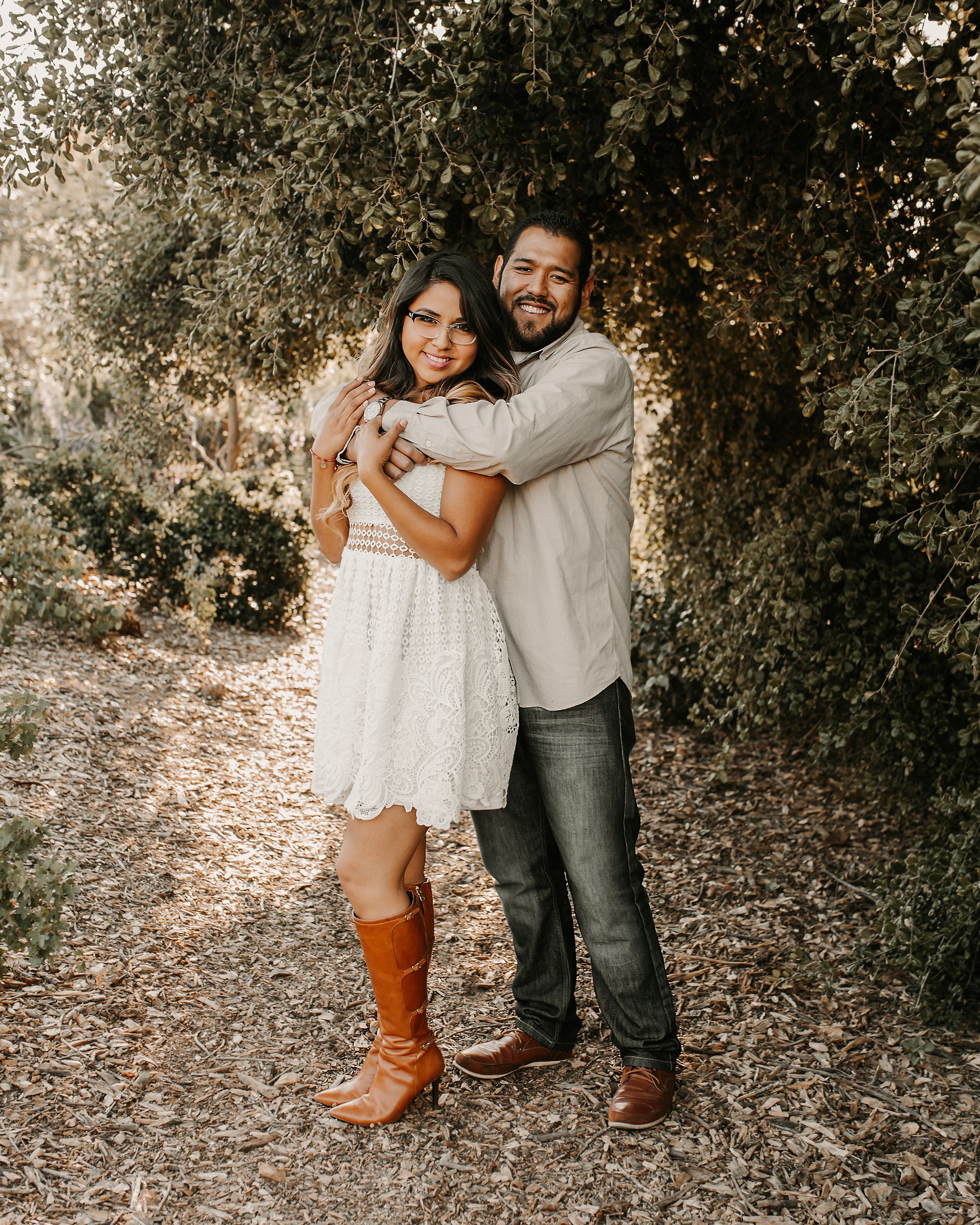Couple in casual attire poses for romantic engagement photos in a wooded outdoor setting.