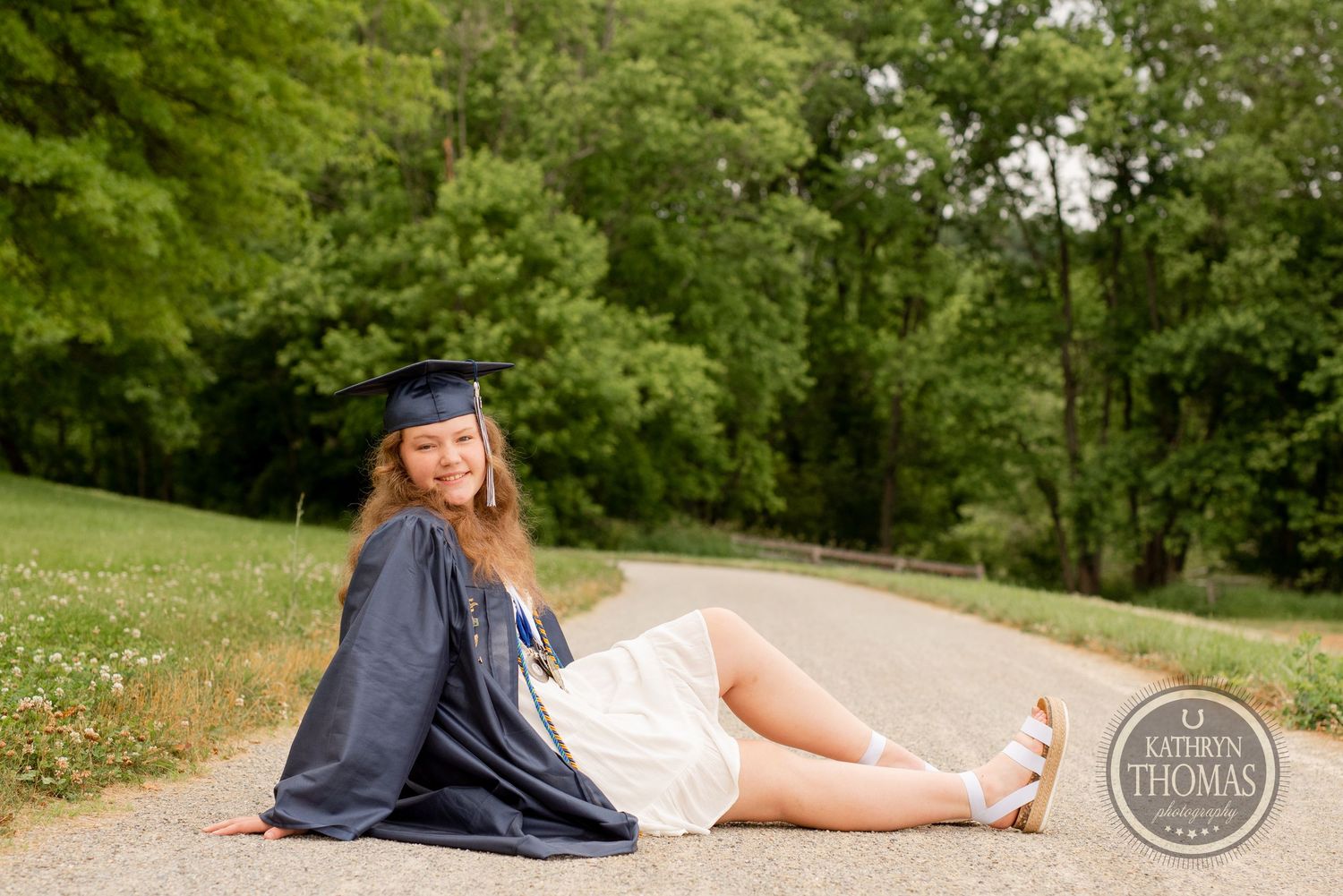 Graduate in cap and gown reclines on path surrounded by green landscape.