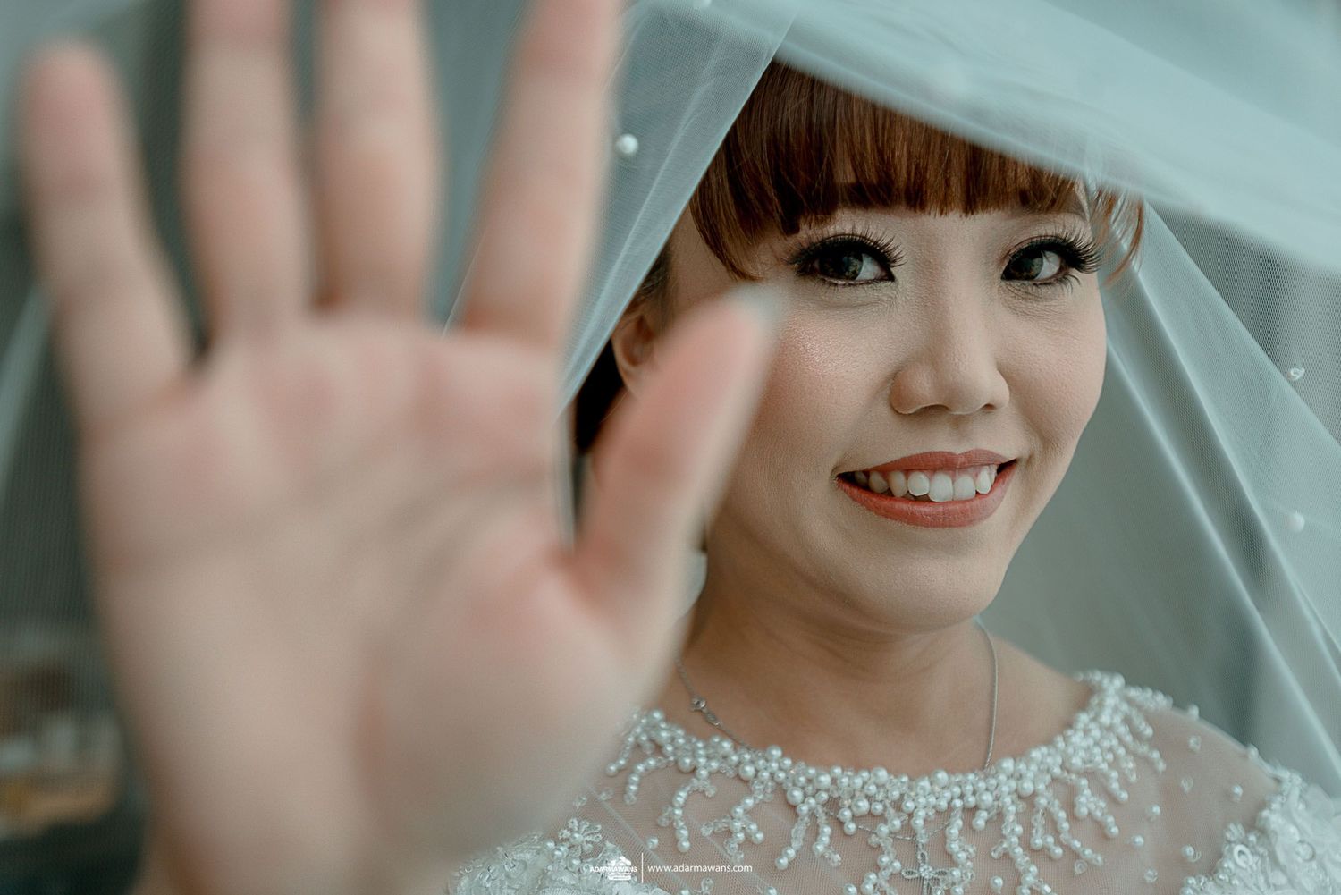 A bride with bangs smiles and waves at the camera through her delicate veil.