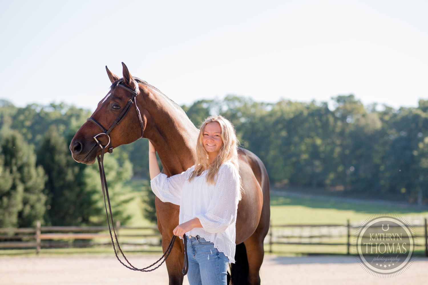 Woman in white shirt and jeans standing with bay horse in sunny outdoor paddock.