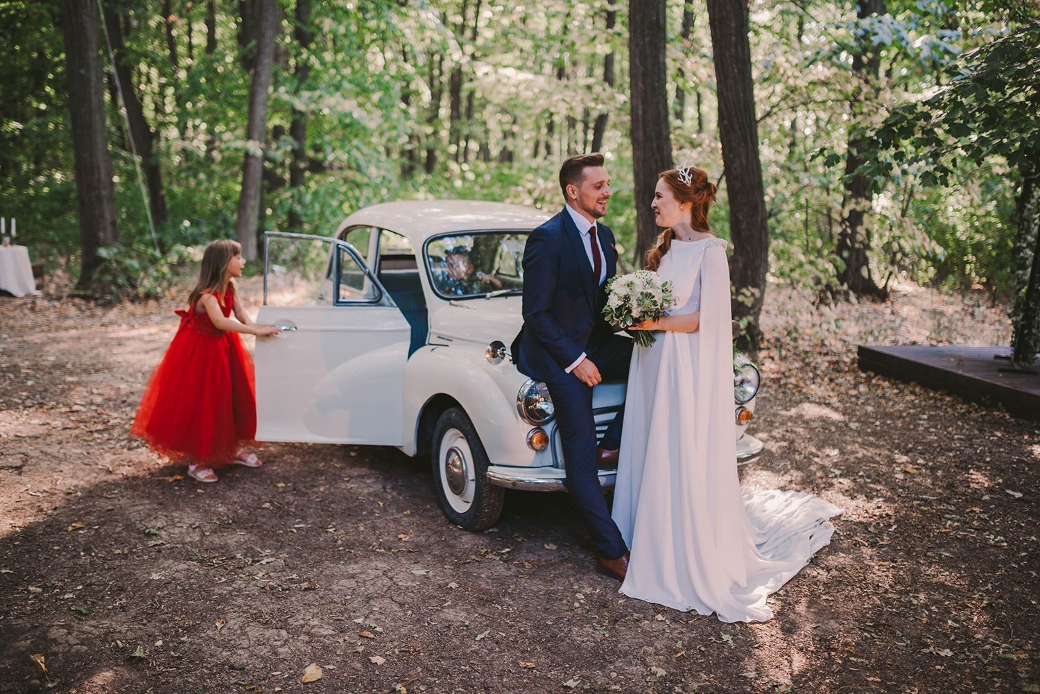 A vintage white car serves as a backdrop for outdoor wedding photos in a forest.