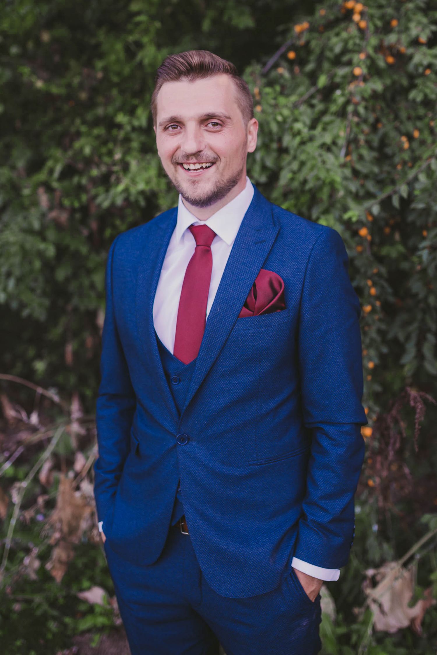 Groom in navy blue suit with red tie poses outdoors against natural foliage background.