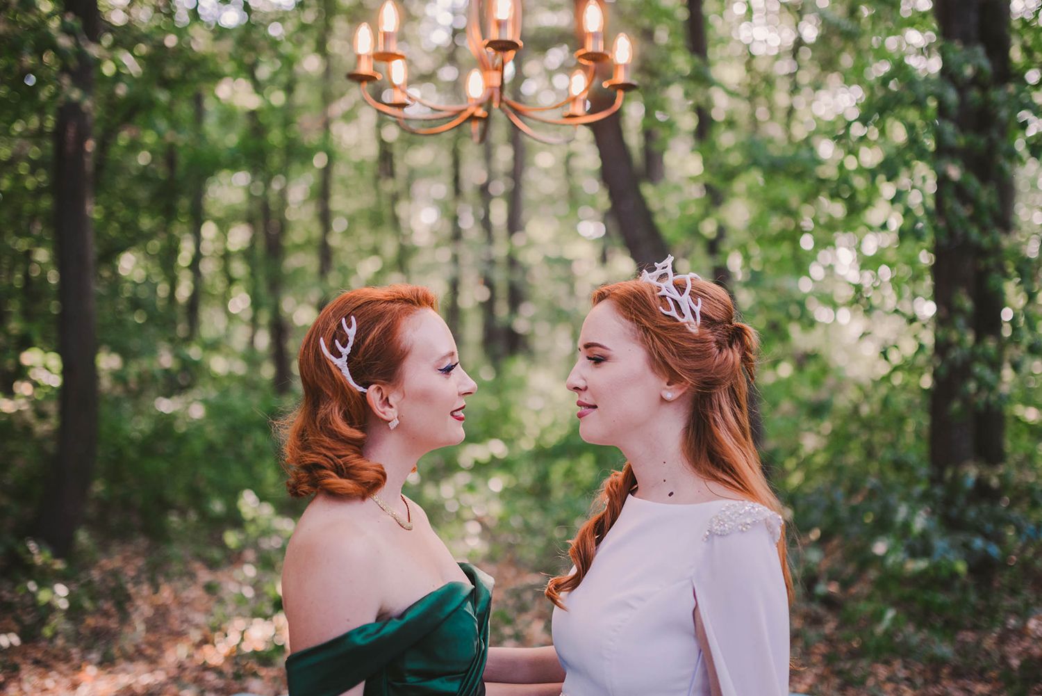 Two women in contrasting white and green formal dresses under chandelier in forest setting.