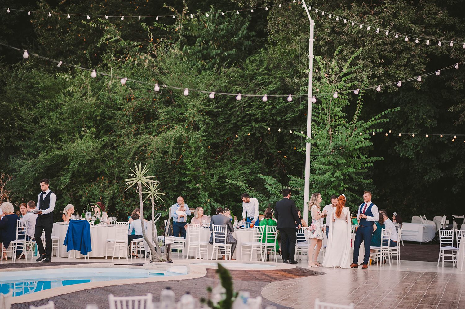 Outdoor wedding reception with string lights and white chairs in garden setting.