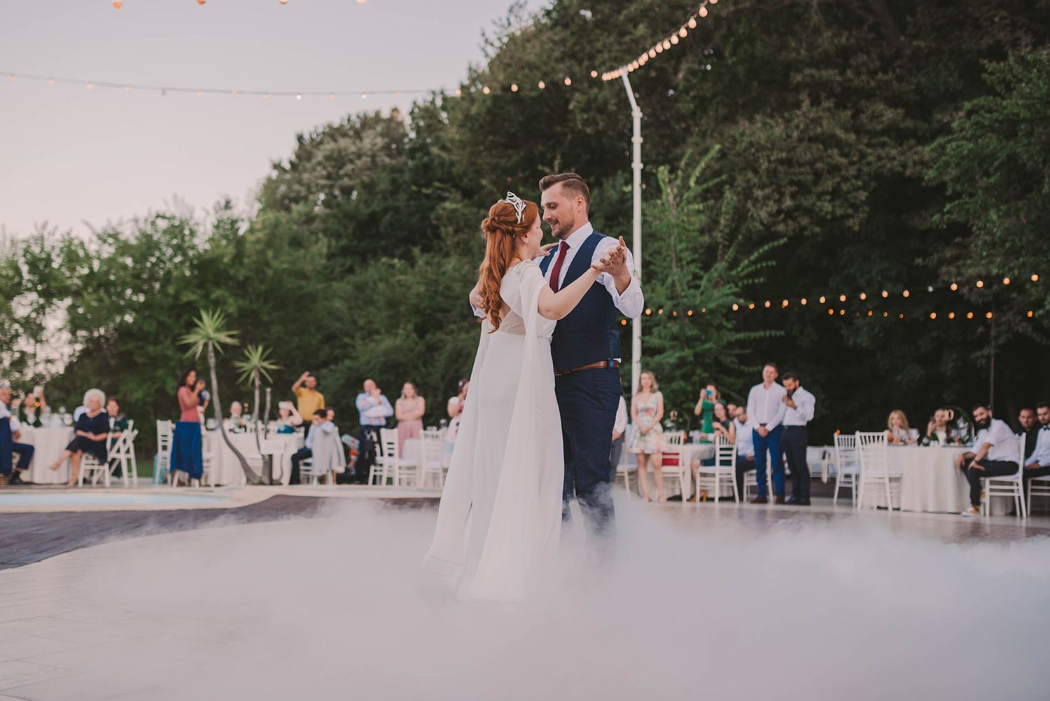 Couple sharing first dance on outdoor dance floor under string lights at dusk.