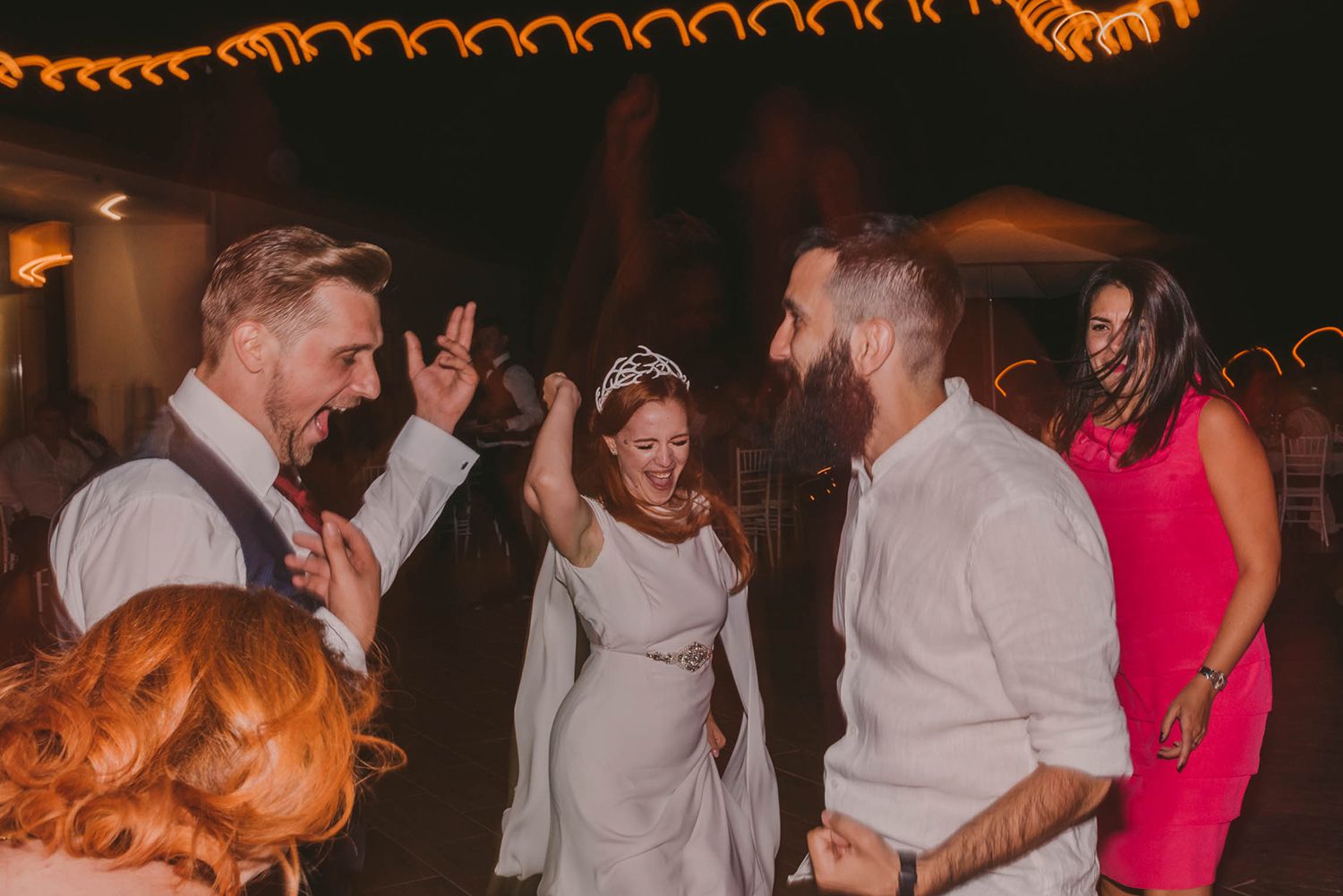 Wedding guests dance and celebrate under warm string lights at evening reception.