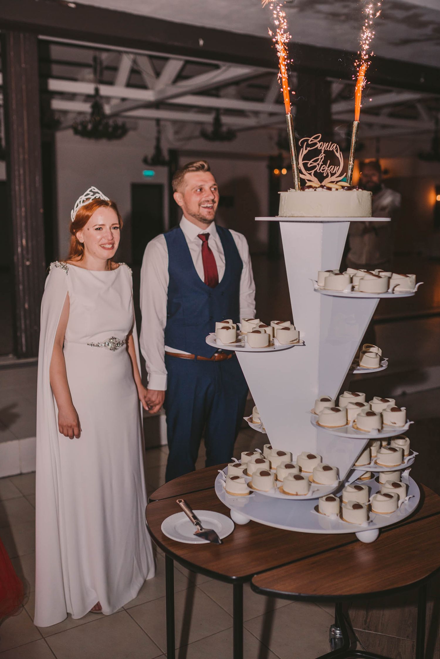 Wedding couple stands beside an elegant three-tiered white wedding cake display at their reception.