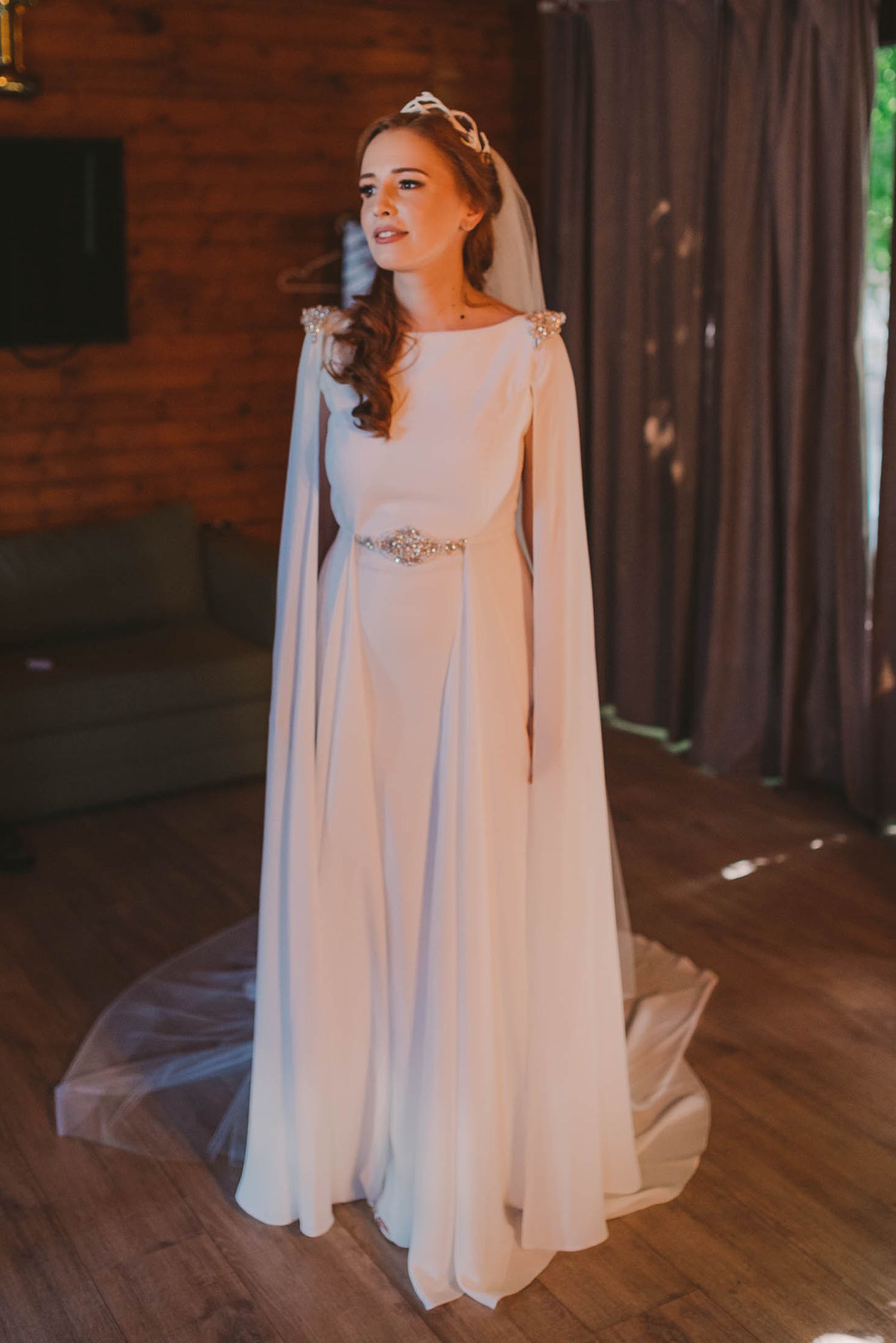 Bride in flowing white cape dress poses in rustic wooden interior setting.
