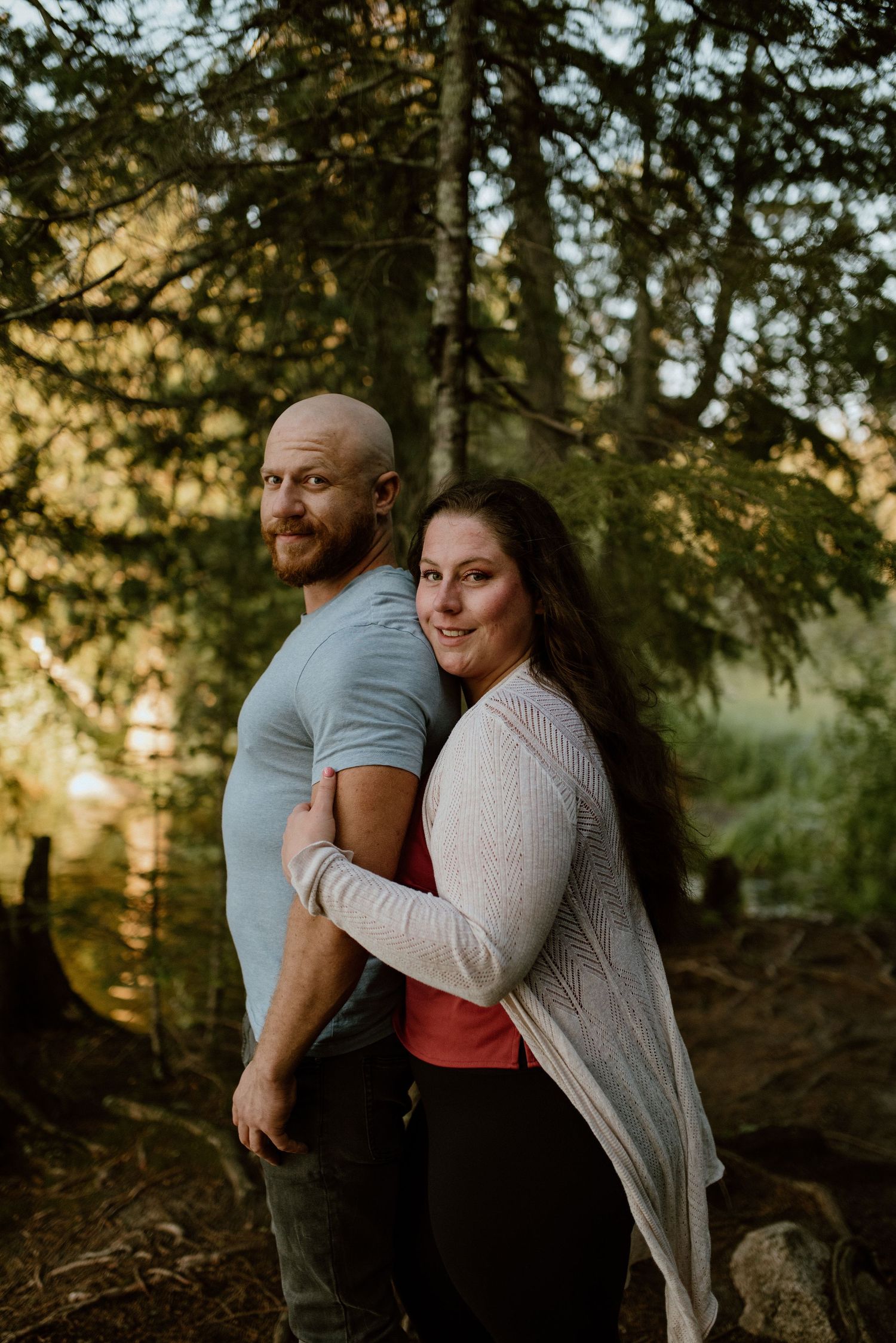 Couple poses together in a sunlit forest setting wearing casual attire.