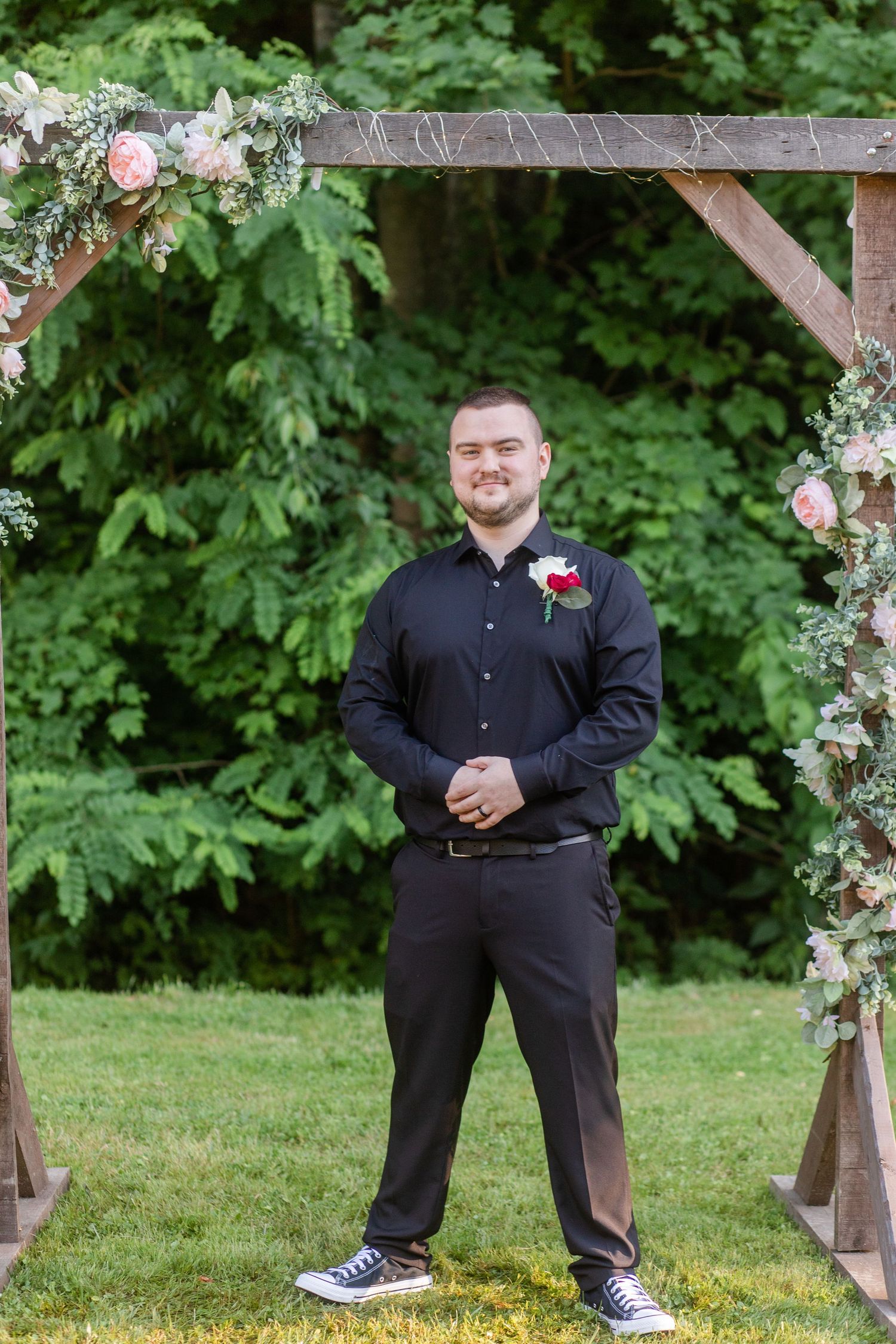 Groomsmen in black suits and sneakers posing under floral arch at outdoor wedding.