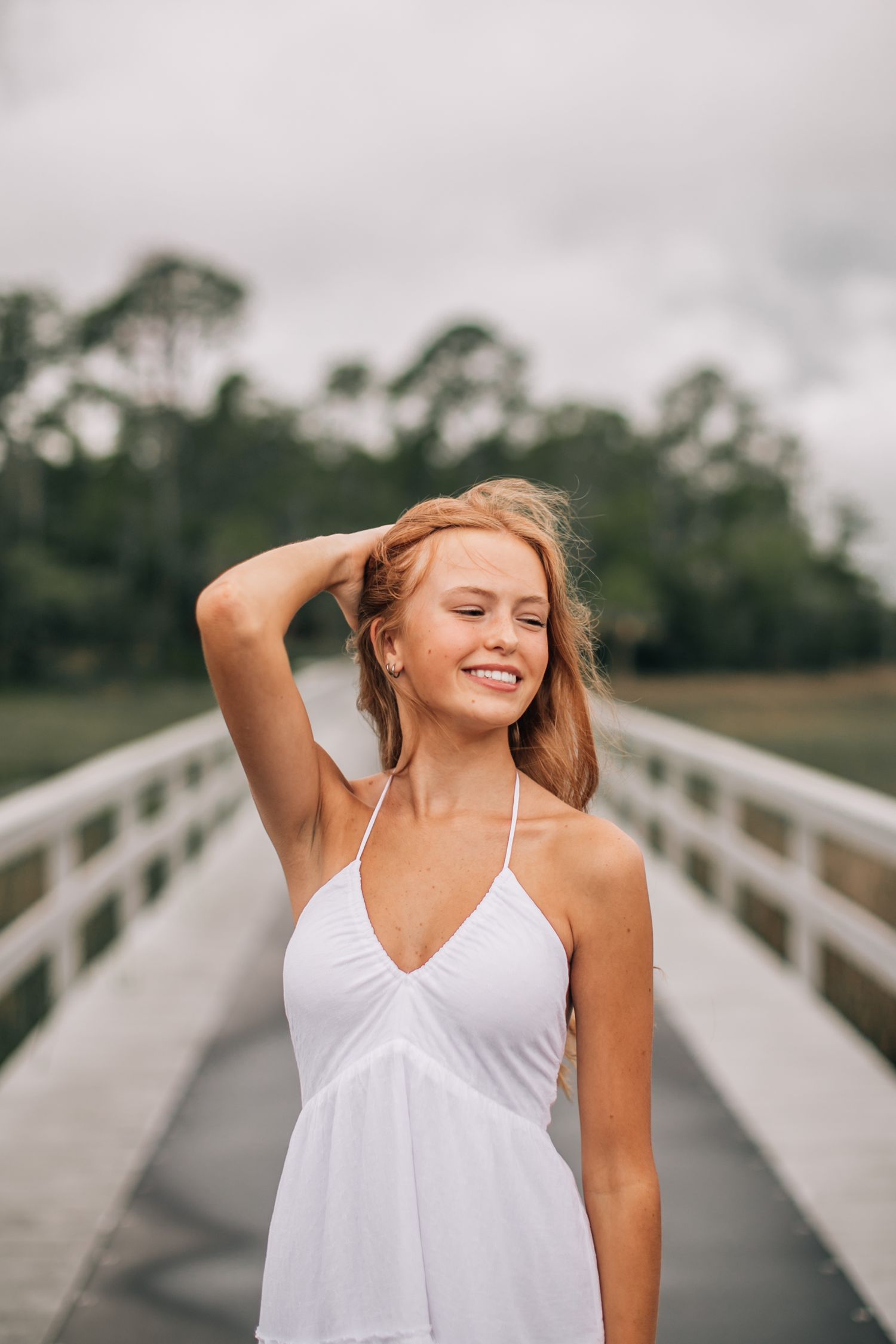 Person in white dress smiling on wooden boardwalk with natural scenery.