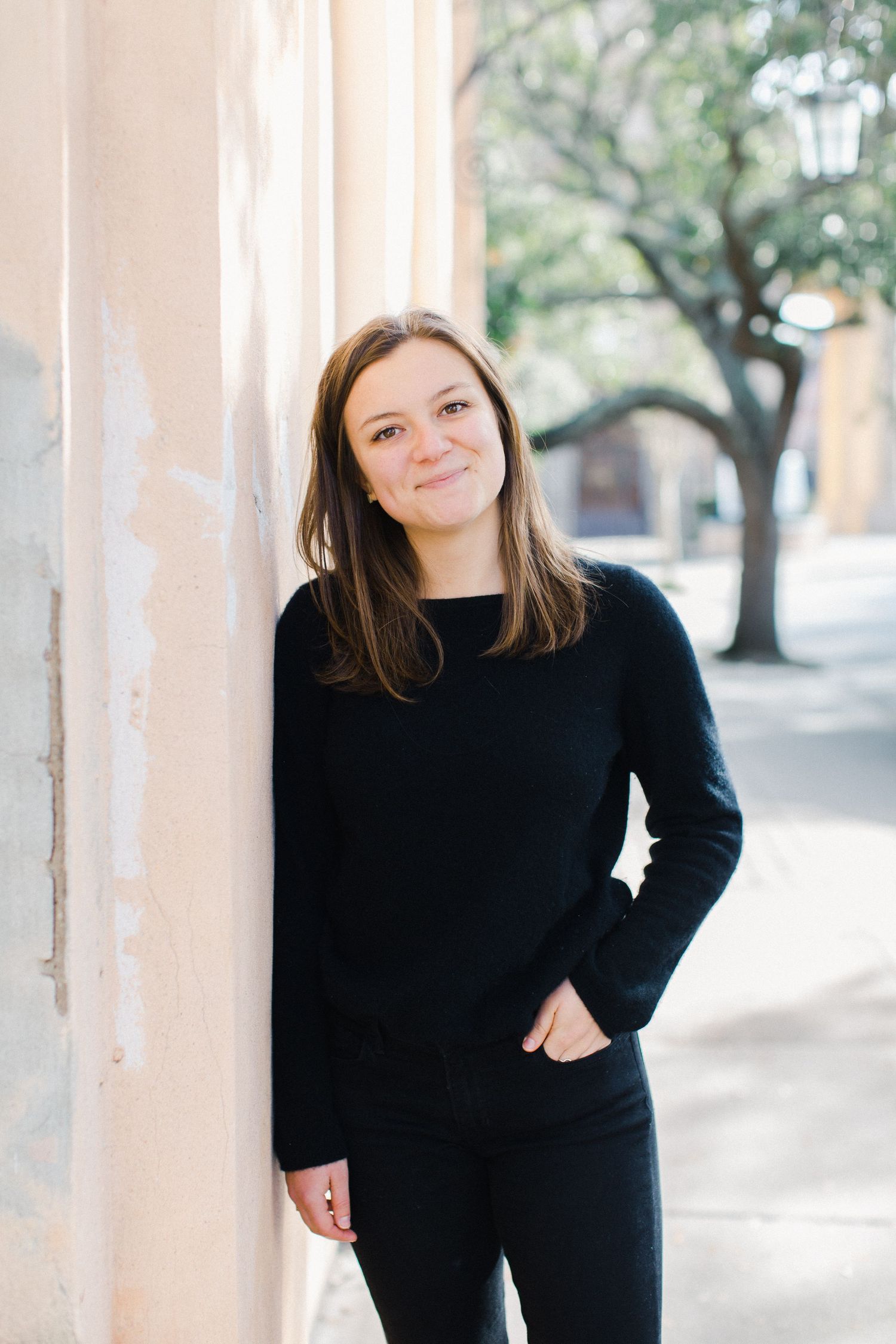Graduating college student in black sweater poses casually against light colored wall outdoors at College of Charleston for senior photo.
