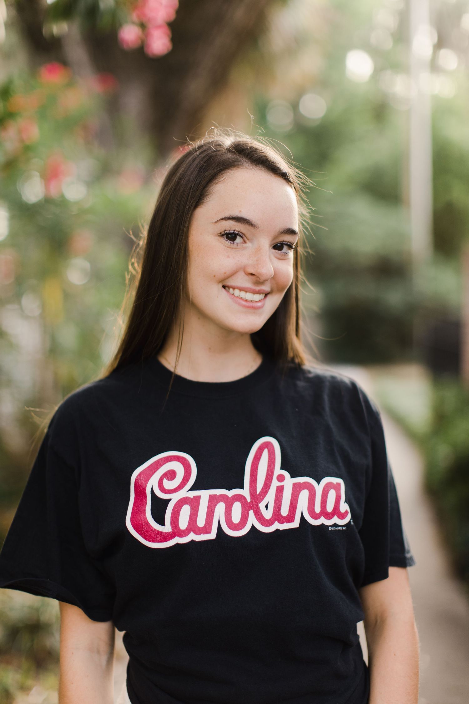 High school graduating student wearing black Carolina t-shirt smiles outdoors with blurred floral background posing in senior photos.
