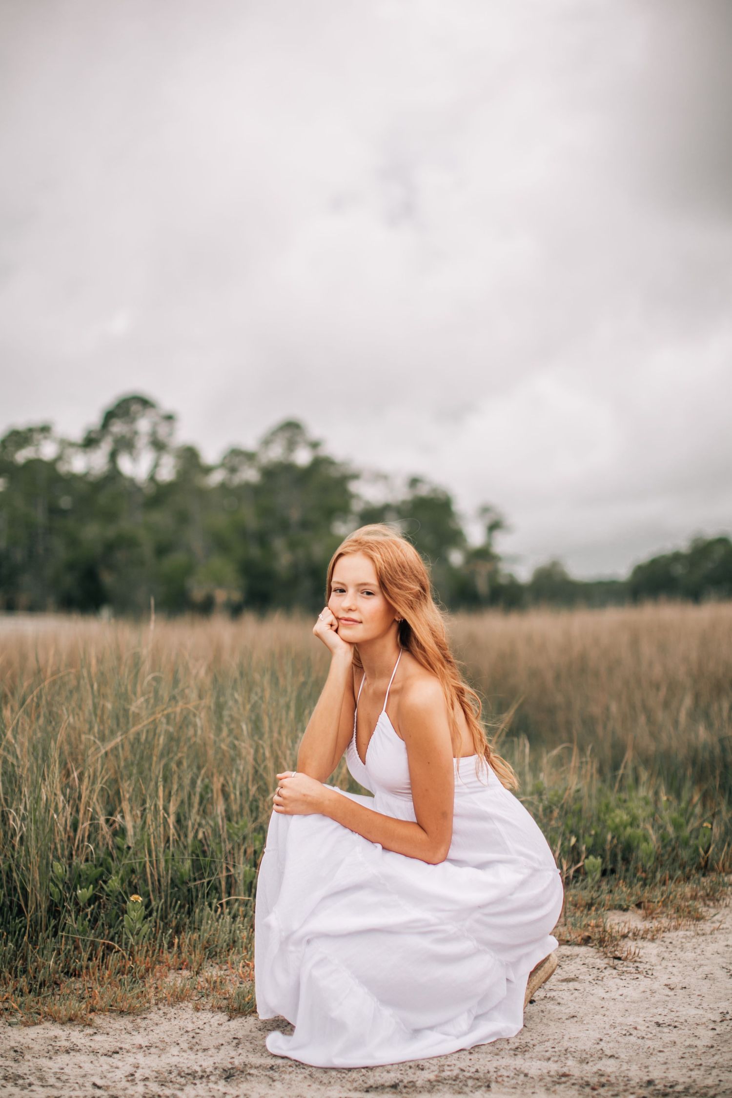Person in white dress sitting near marsh with trees in background.