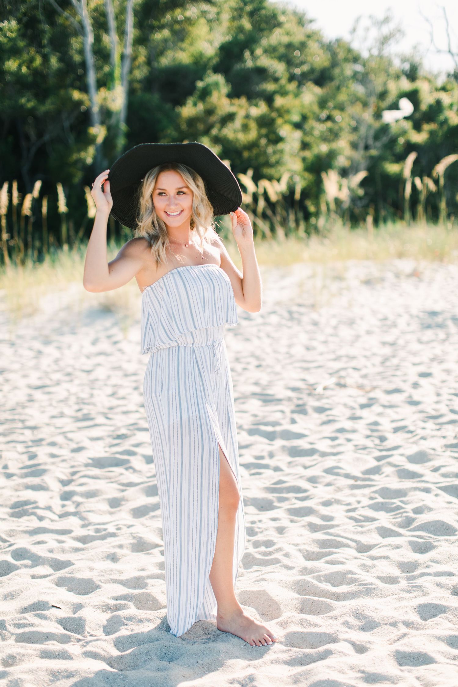 Senior session girl dressed in striped maxi dress poses playfully with black hat on sandy beach in Hilton Head, SC.