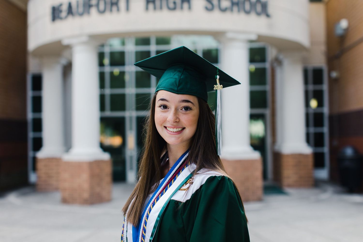Graduate in dark green cap and gown poses in front of Beaufort High School entrance for senior photography session.