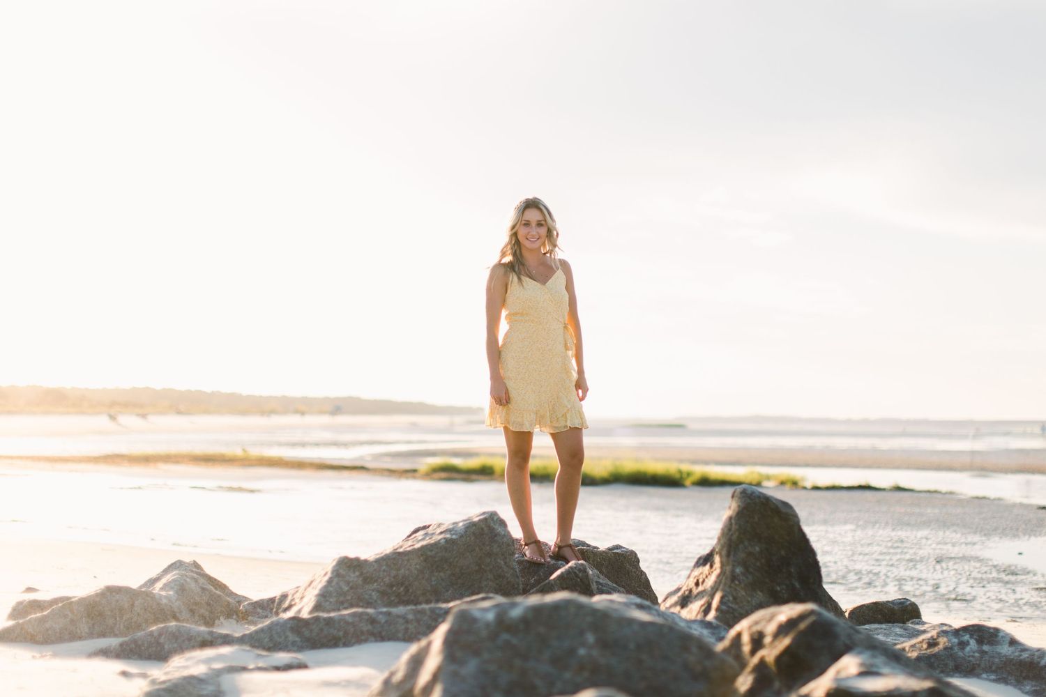 Woman in beige dress stands on coastal rocks during golden sunset.