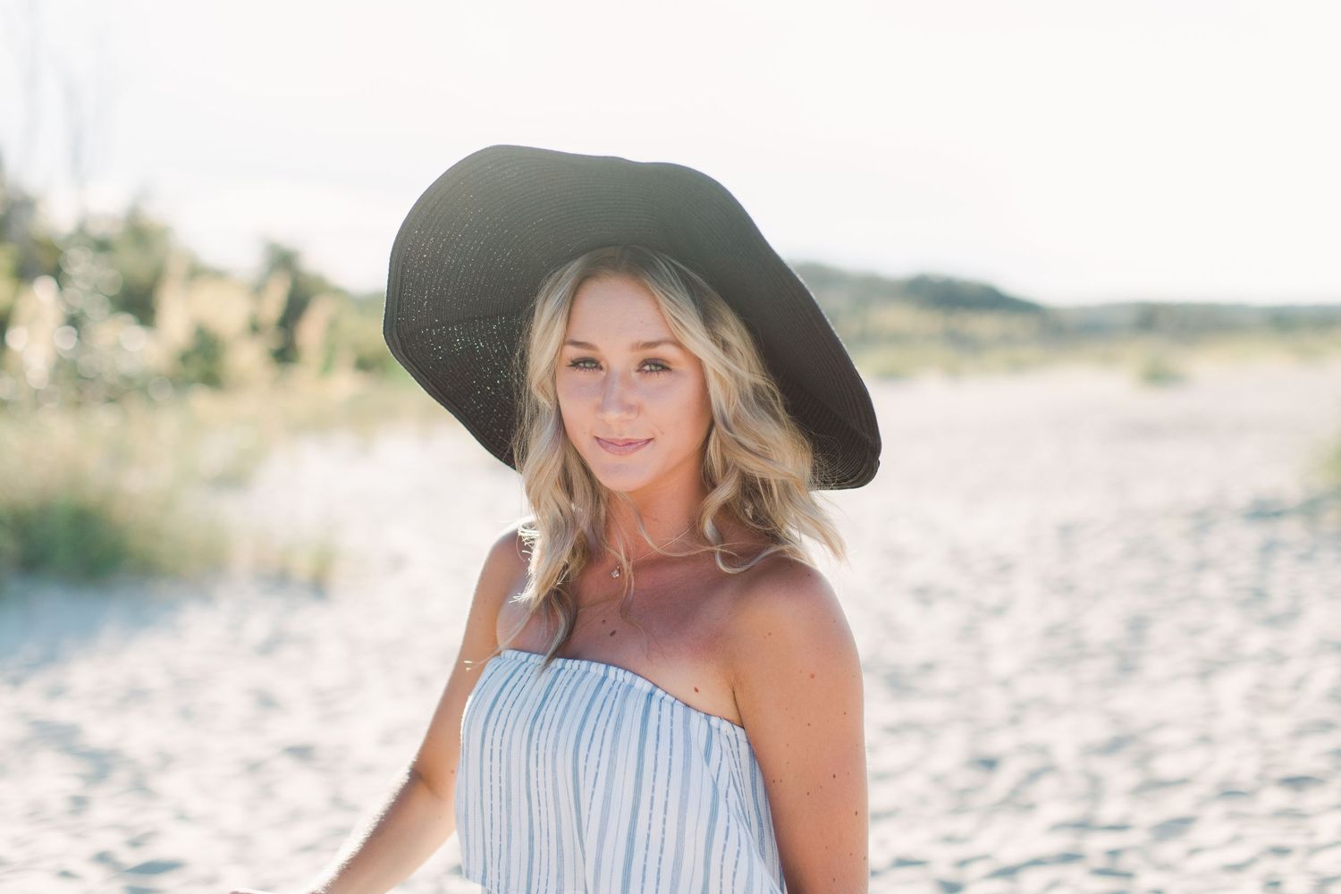 Person in striped dress and wide-brim hat standing on sunny beach.