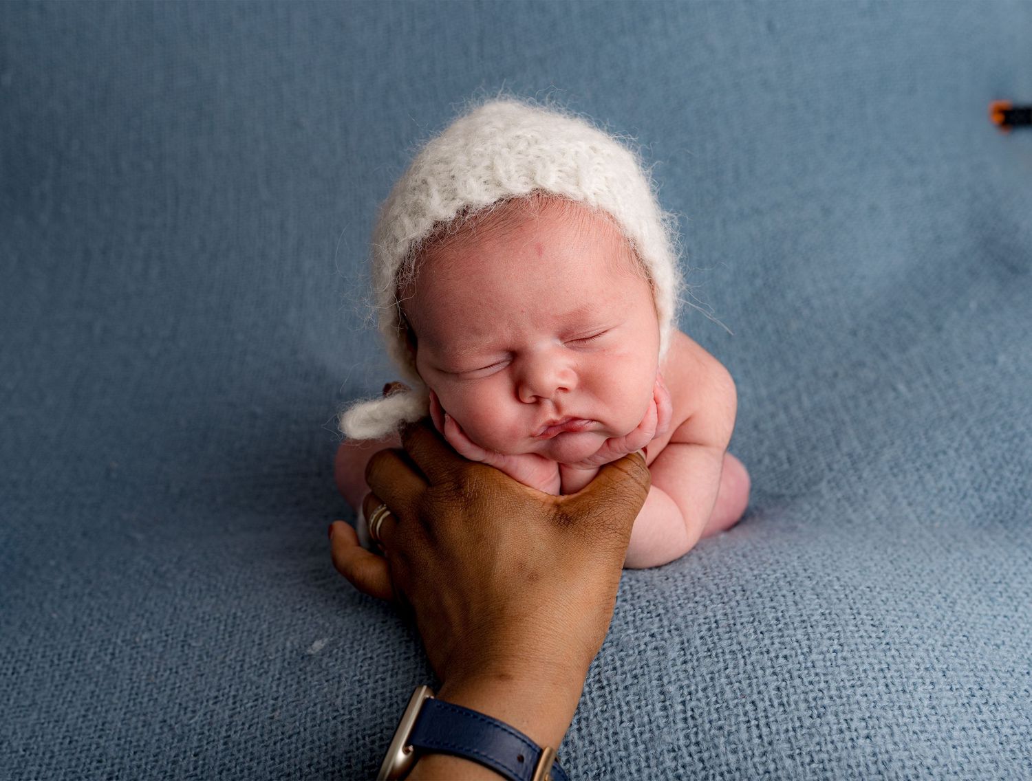 A froggy composition of a sweet sleeping newborn wearing white bonnet rests on blue backdrop.