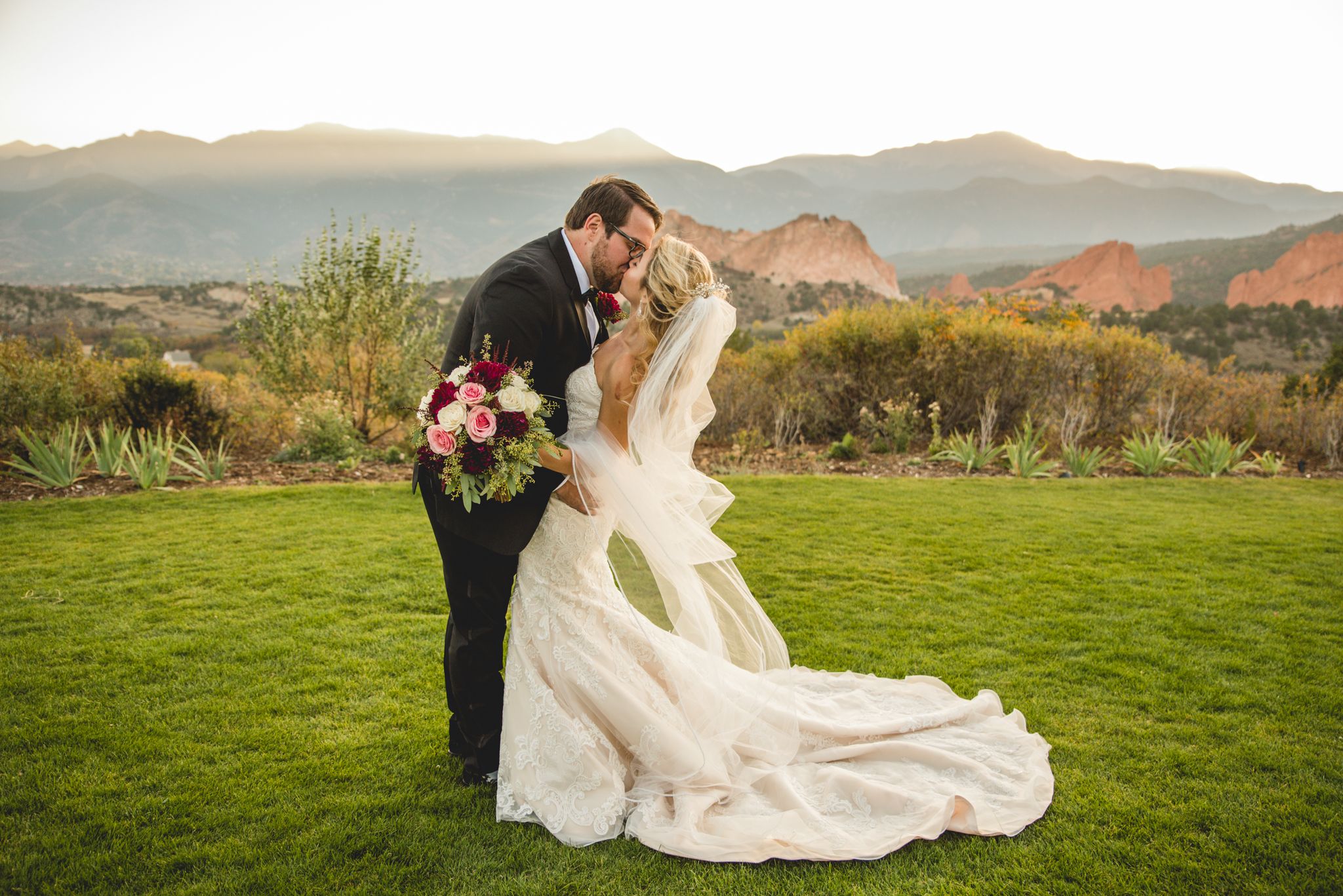 A wedding couple embraces on a hilltop lawn with stunning mountain views and sunset lighting in the background.