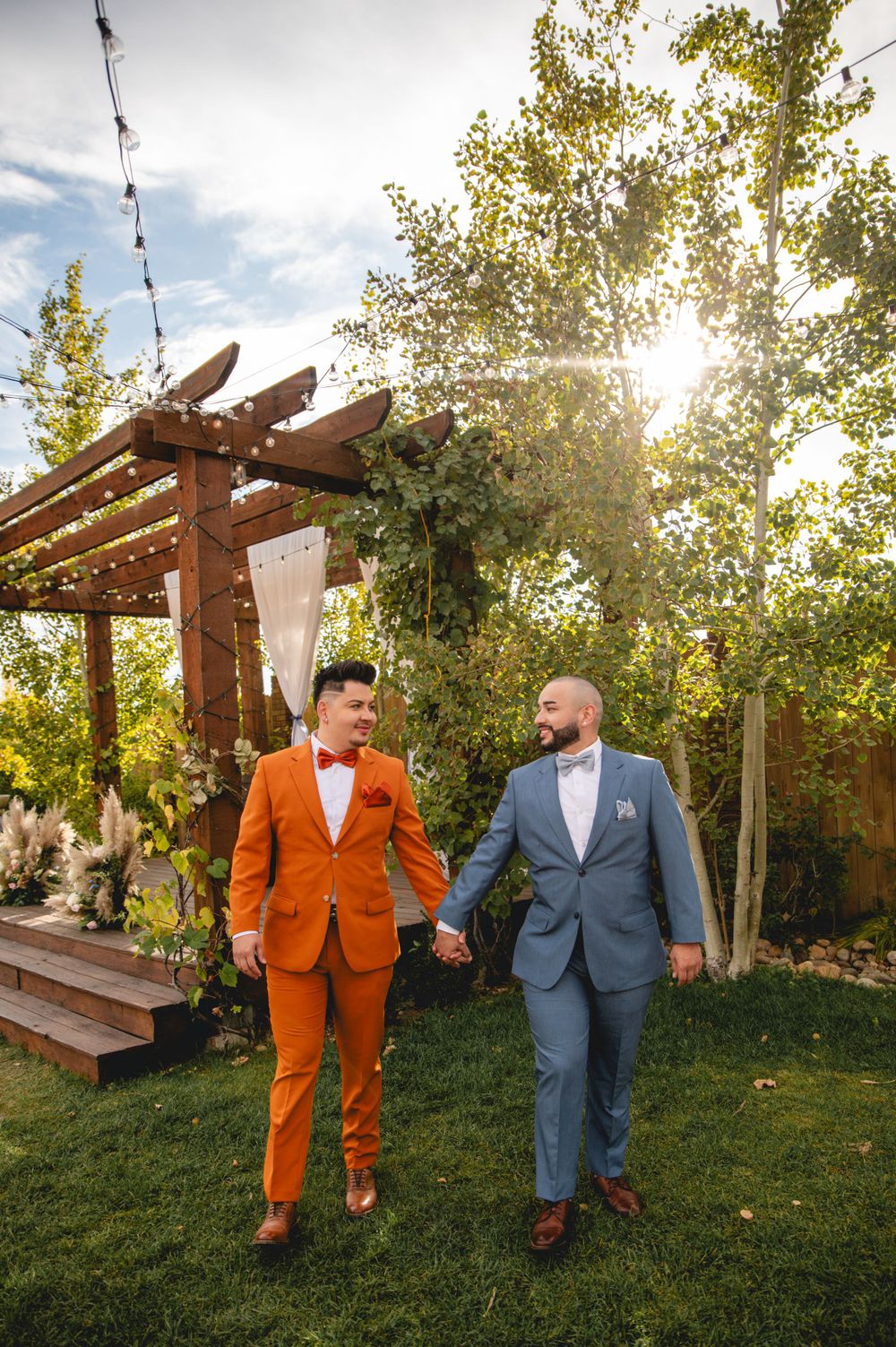 LGBTQ+ couple in formal attire, one in bright orange and one in blue, walking together in a garden with a wooden pergola.