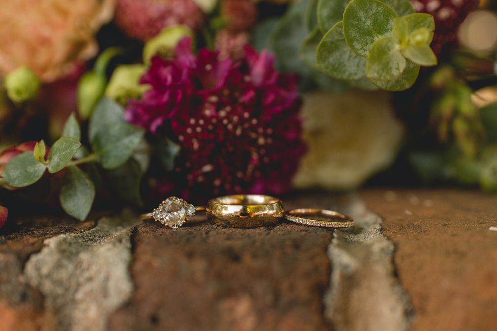 Wedding rings displayed on brick against a background of burgundy flowers and green foliage.