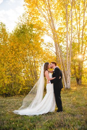 A romantic wedding couple embraces in front of golden autumn trees at sunset with a long flowing veil trailing behind.