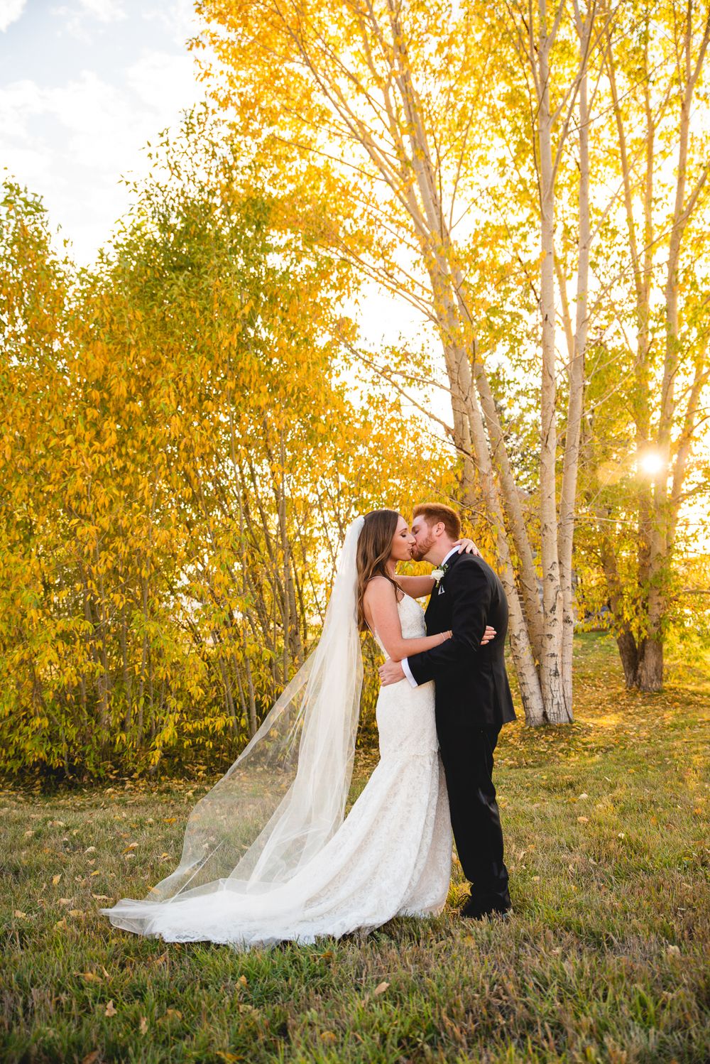A romantic wedding couple embraces in front of golden autumn trees at sunset with a long flowing veil trailing behind.