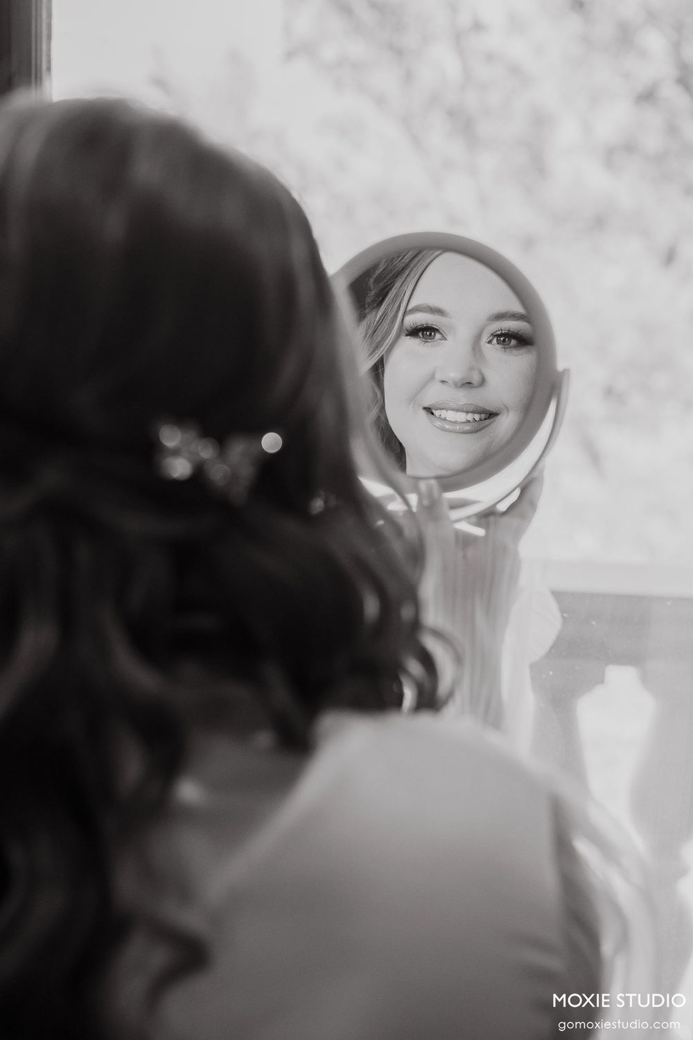 Black and white reflection in mirror shows a smiling person with styled hair and makeup looking at their own image.