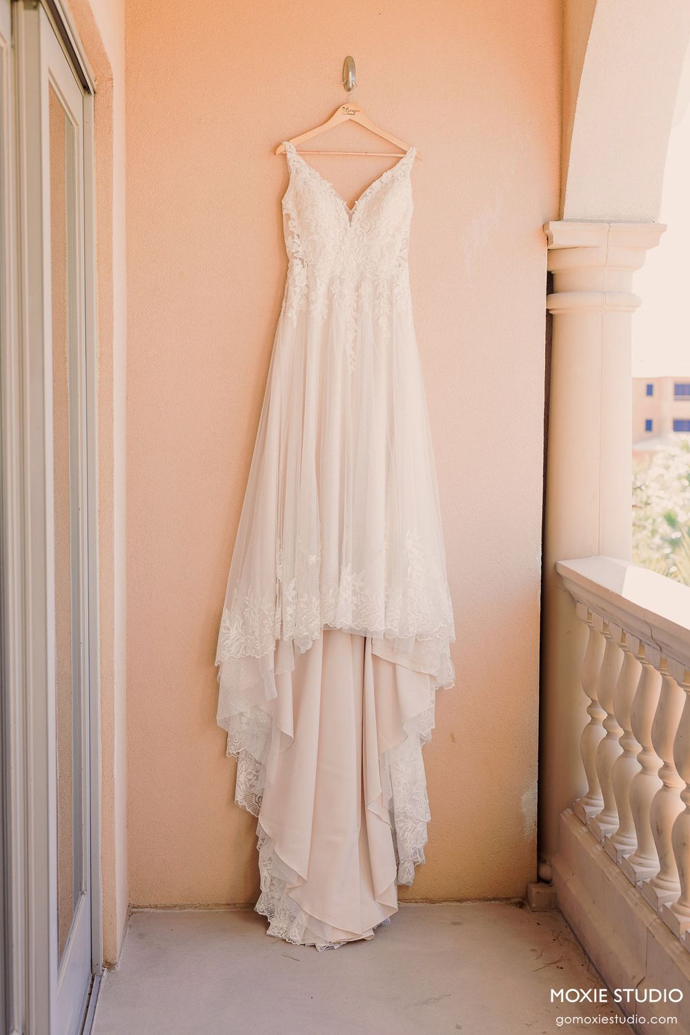 Elegant white lace wedding dress hanging on display in a sunlit balcony with cream colored walls.