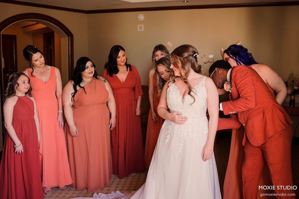 Bridesmaids in coral dresses stand in line with bride in white gown at indoor wedding venue.