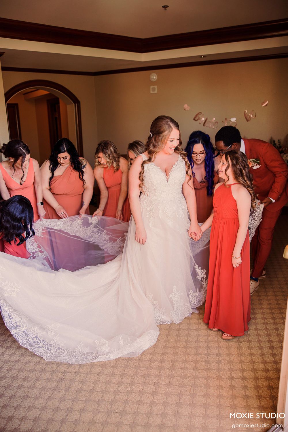 Bridesmaids in coral dresses gather around bride in white lace gown during wedding celebration.
