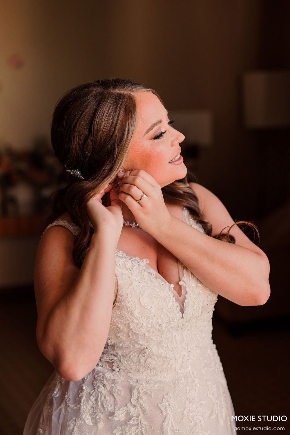 Side profile of bride in white lace gown adjusting her jewelry with elegant hair styling.