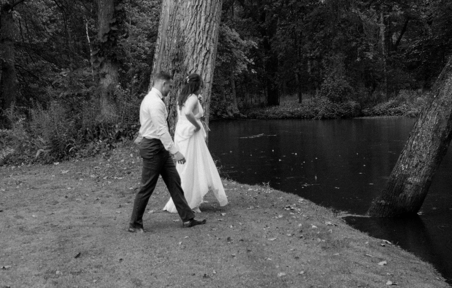 Couple walking along wooded path near water in wedding attire.