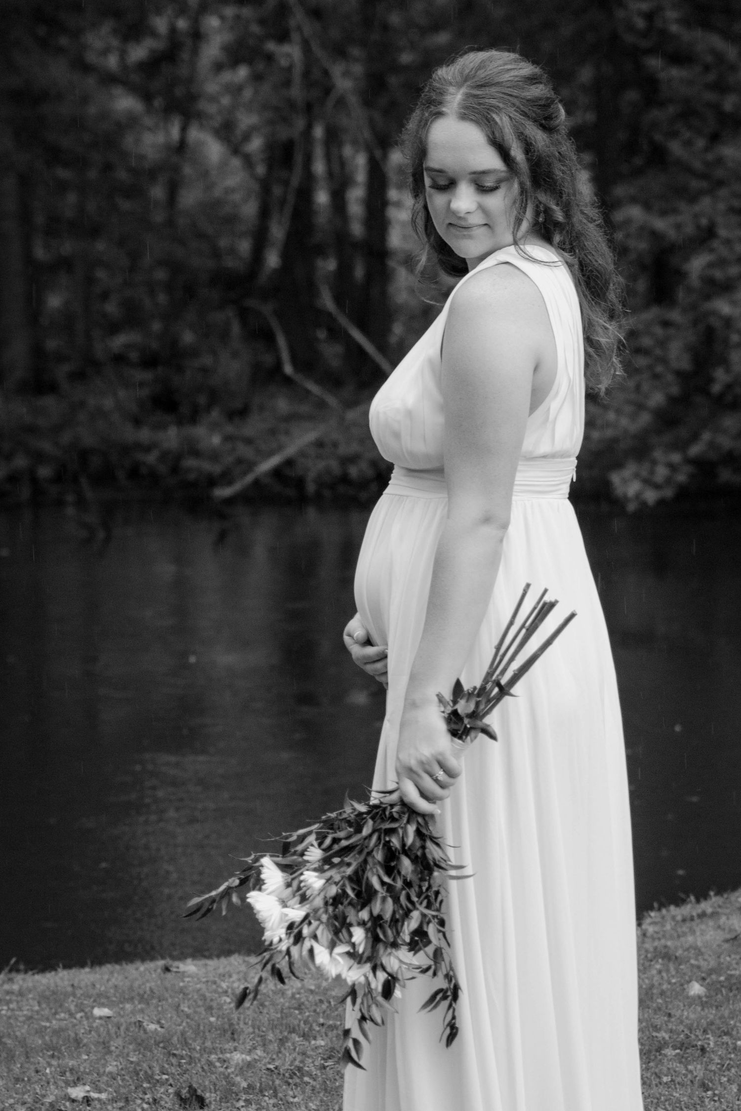 Person in white dress holds bouquet by a serene lake in black and white photography.
