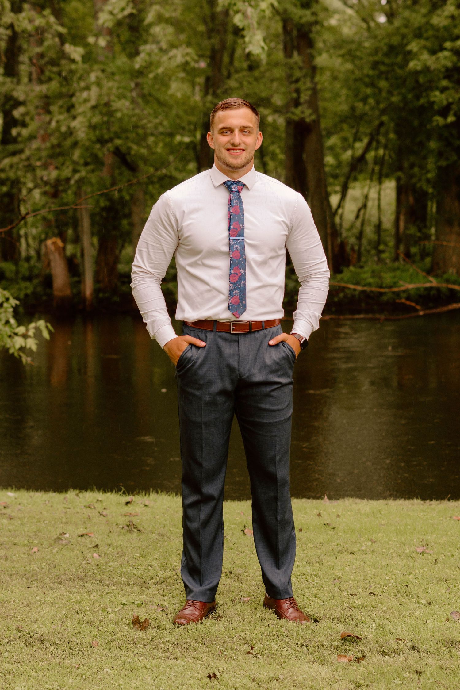 Person in formal attire poses on grass with lake and trees in background.