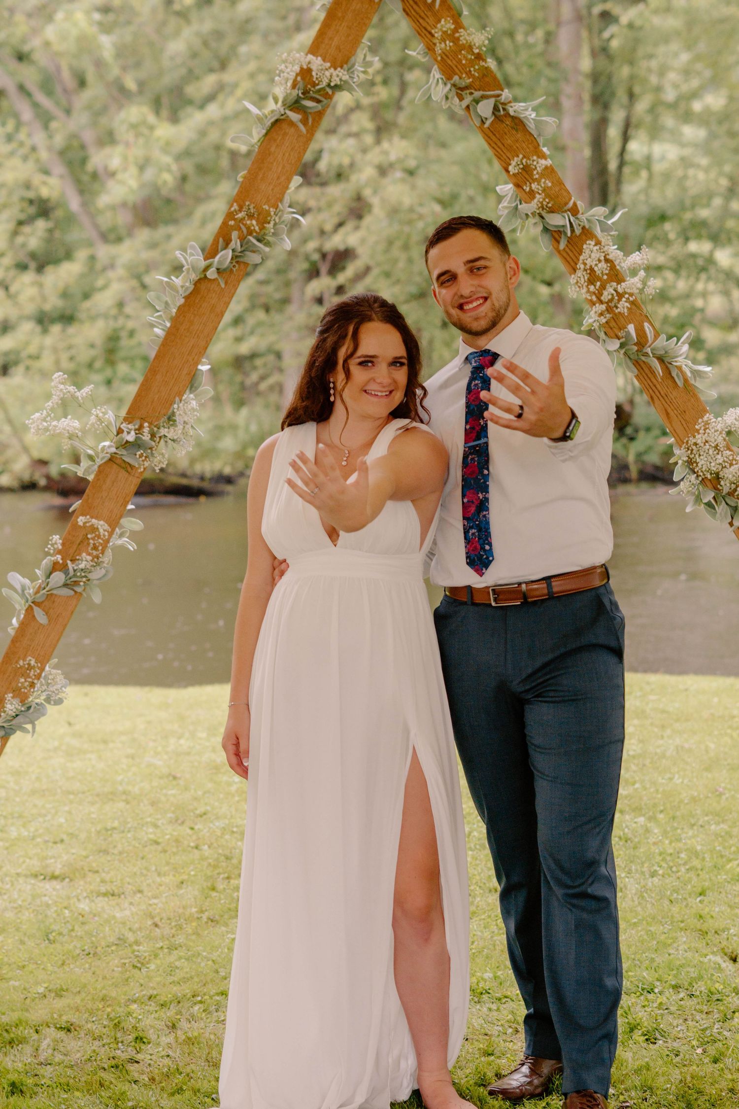 A couple shows their rings under a wooden arch decorated with flowers during an outdoor wedding ceremony.