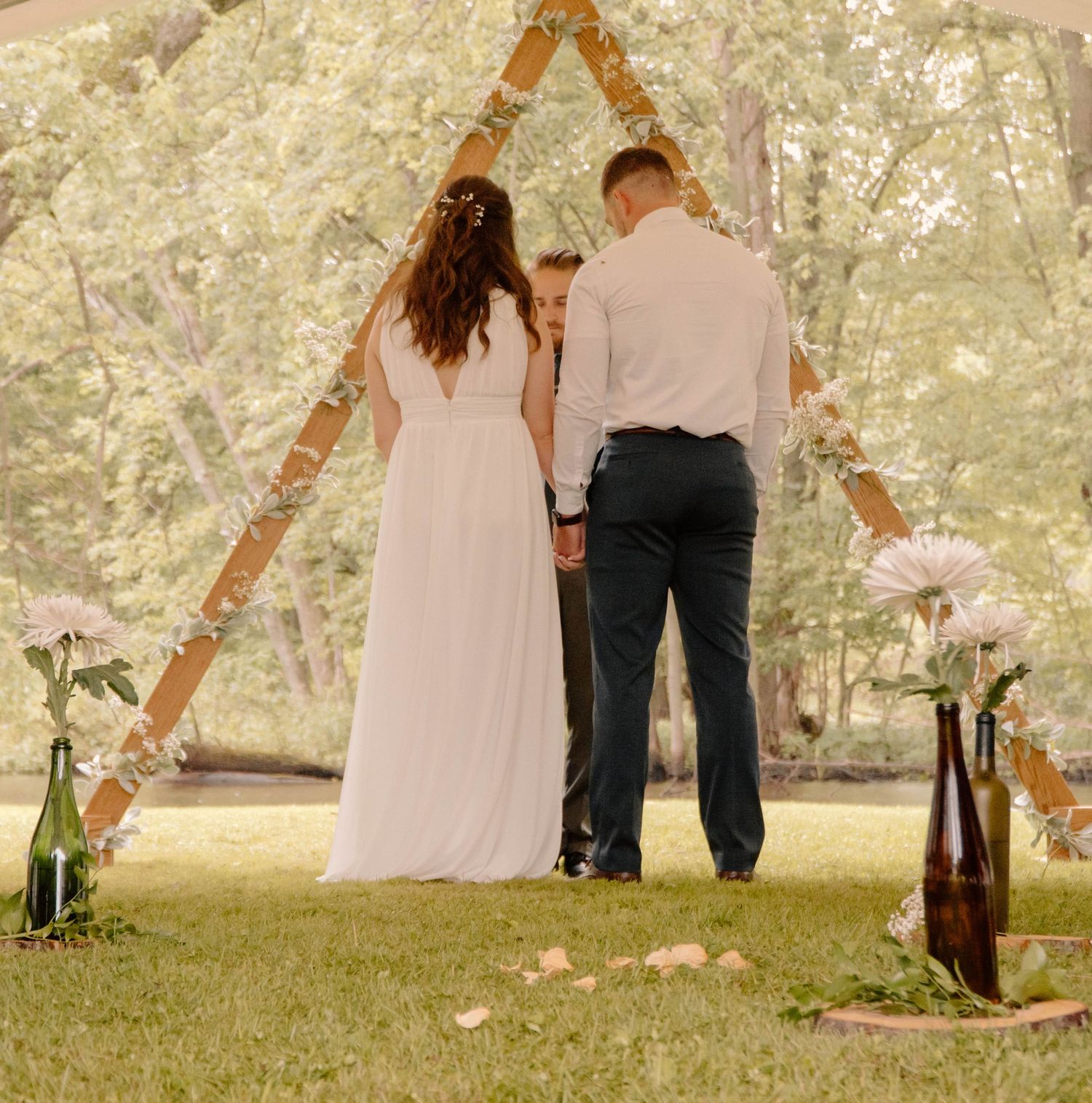 A couple stands beneath a rustic wooden wedding arch decorated with flowers in an outdoor garden setting.