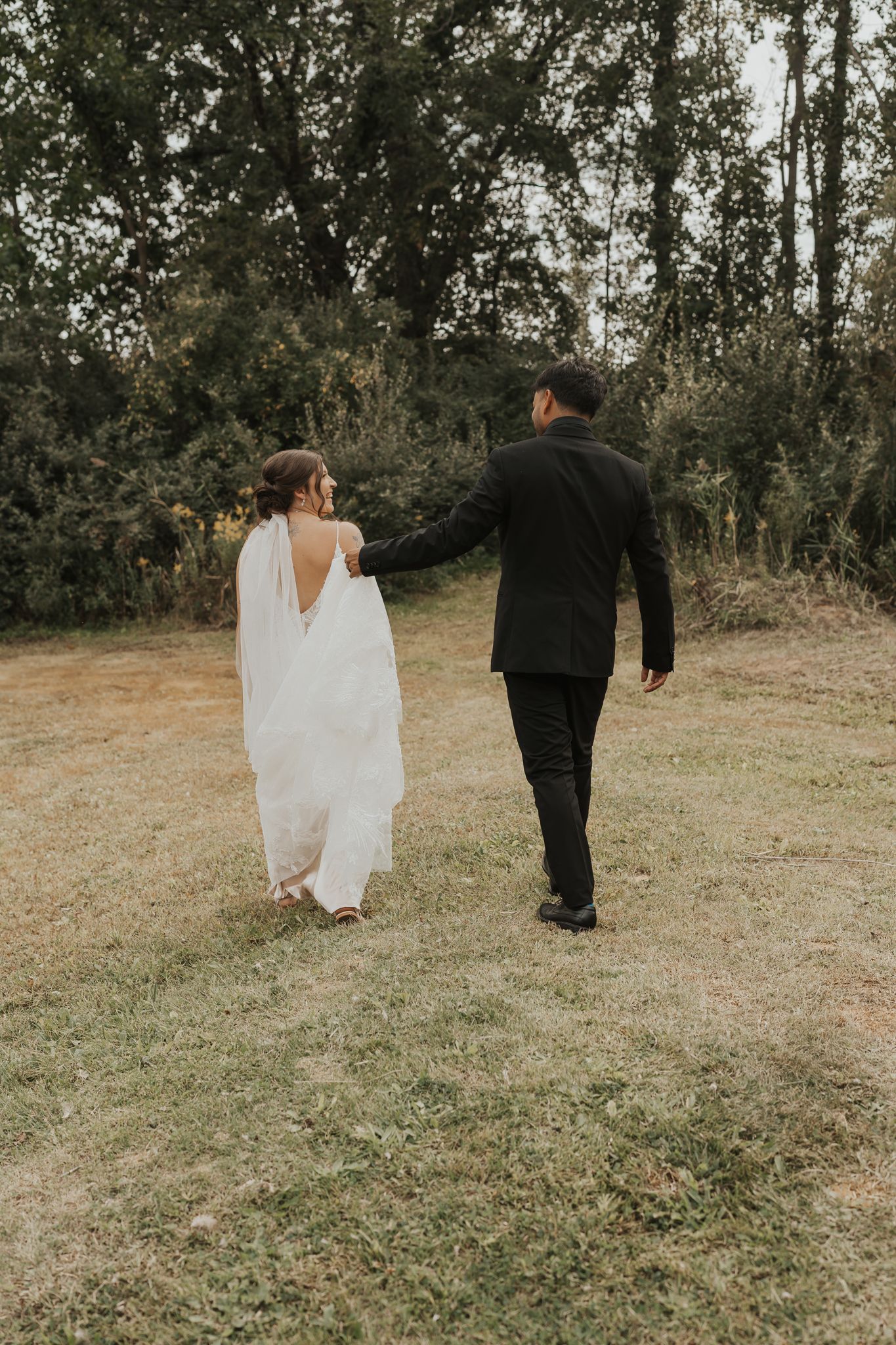 Newlyweds walking hand in hand through a grassy outdoor setting.